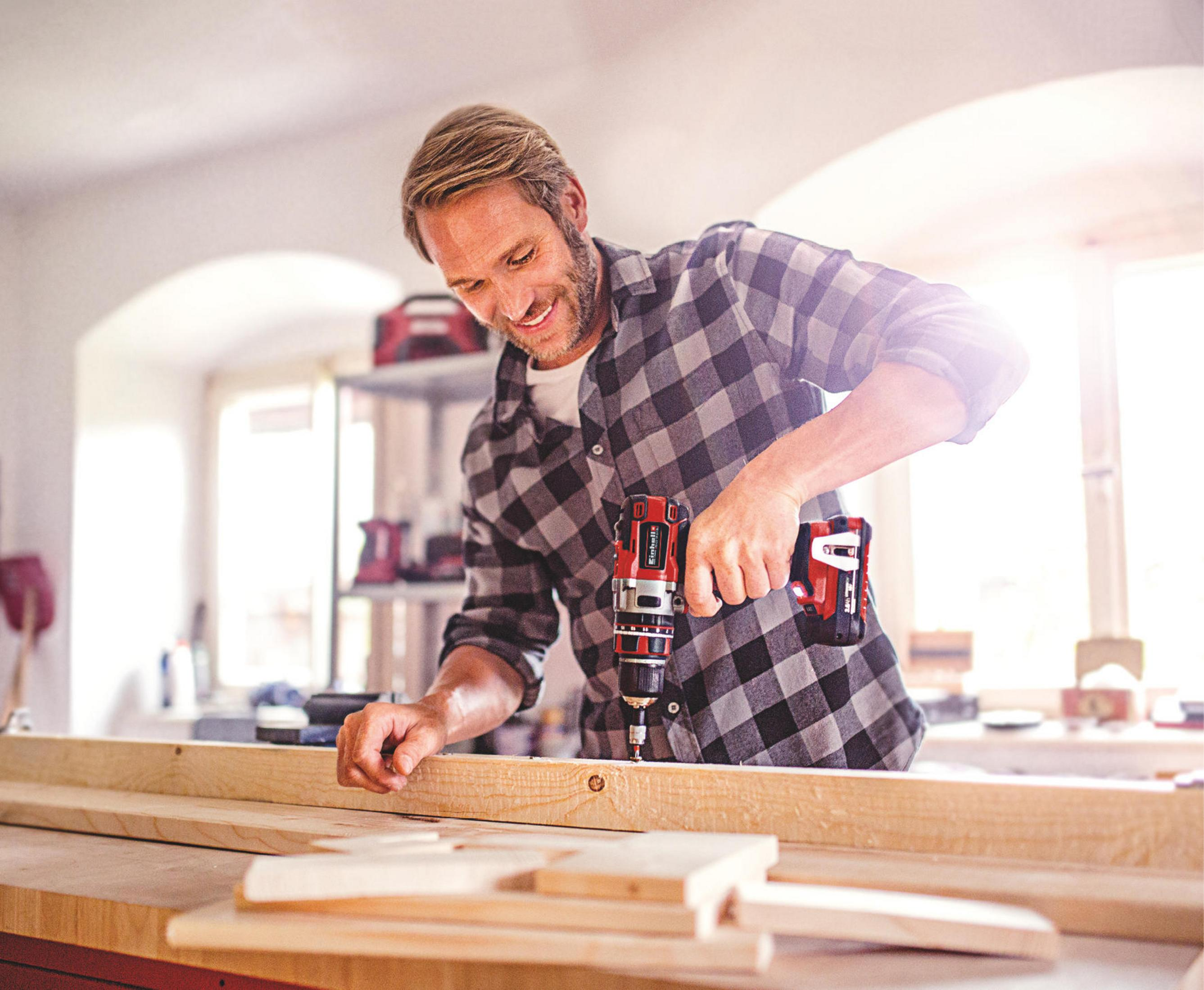 Mężczyzna używający wiertarki na drewnianej desce. W pokoju jest światło słoneczne i inne kawałki drewna.
