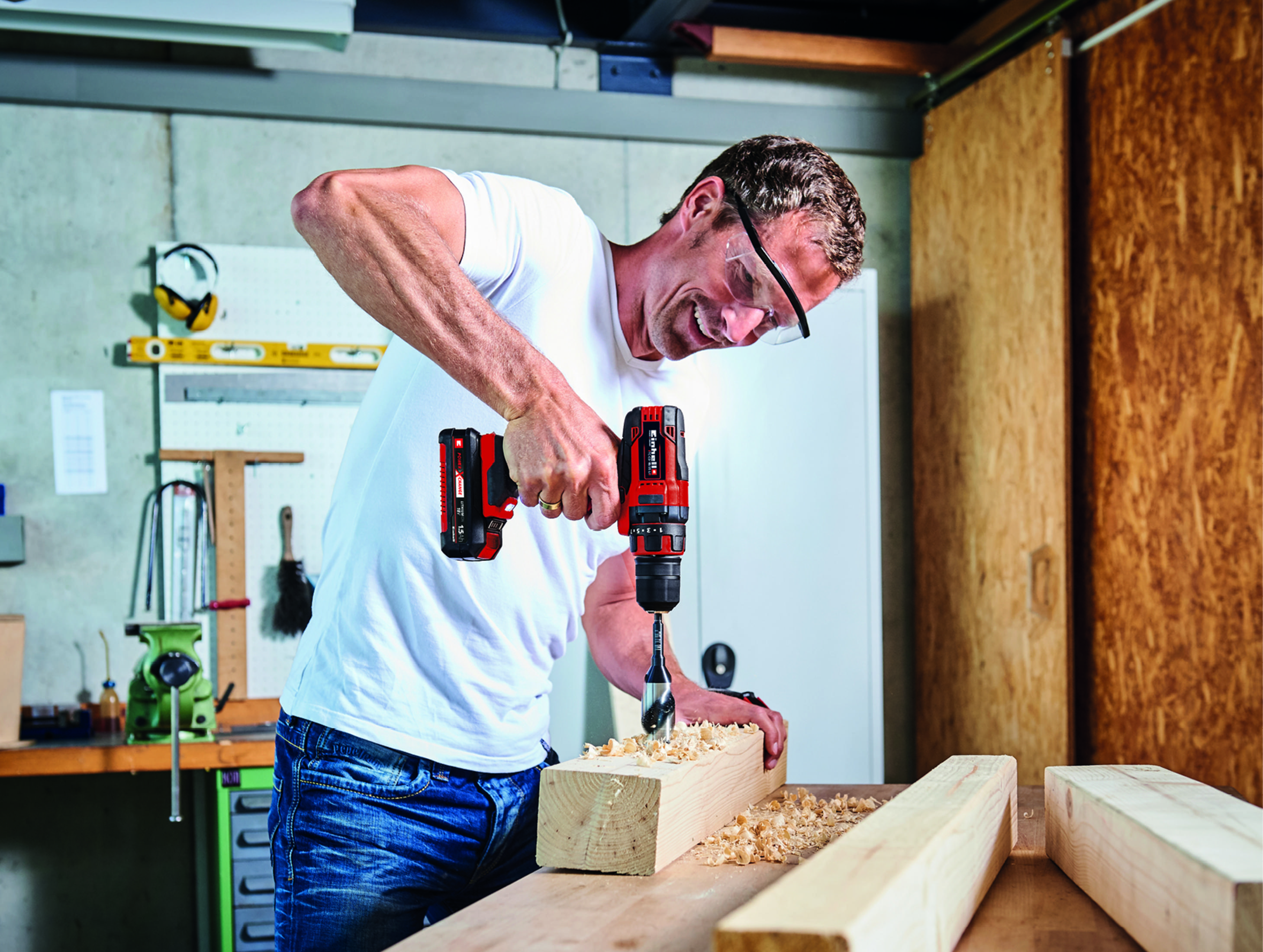 Mężczyzna wierci w drewnie czerwoną i czarną wiertarką akumulatorową. Ma na sobie okulary ochronne.