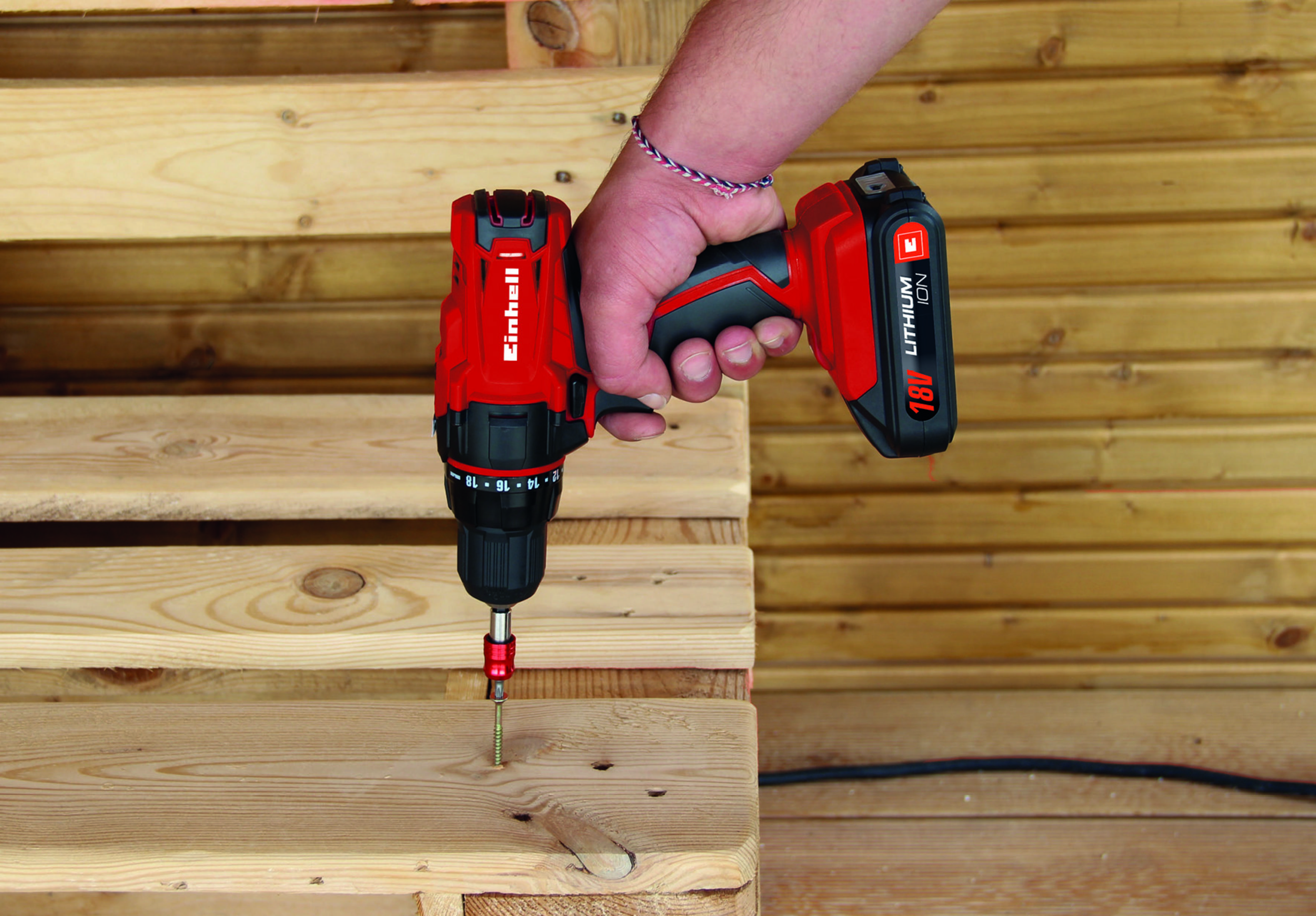 Osoba używająca czerwonej wiertarki do wkręcania w drewnianą deskę. Wiertarka ma czarny uchwyt i jest 18V litowo-jonowa.