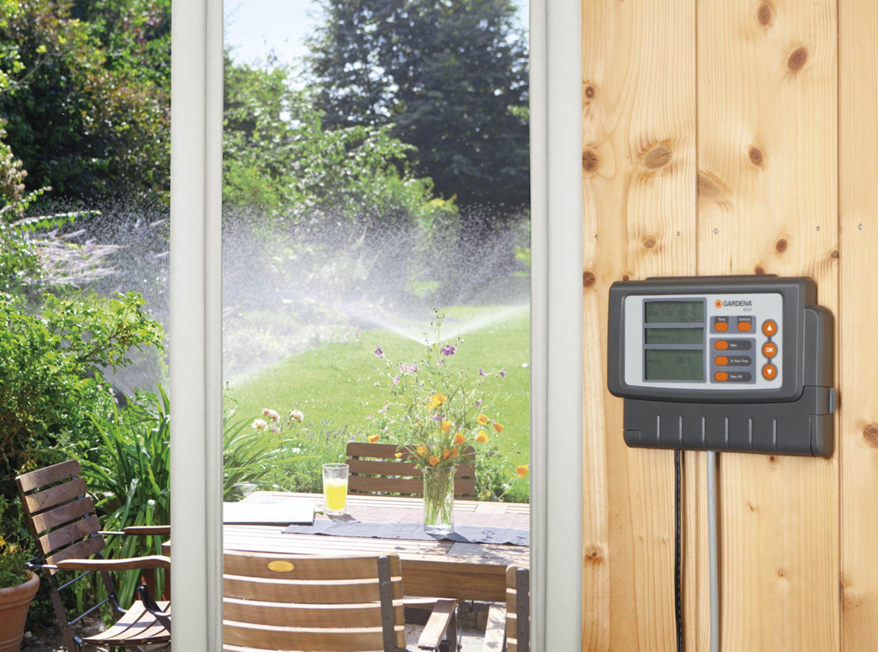 Vista del giardino attraverso una finestra. Un irrigatore innaffia il prato, con un timer su una parete di legno.