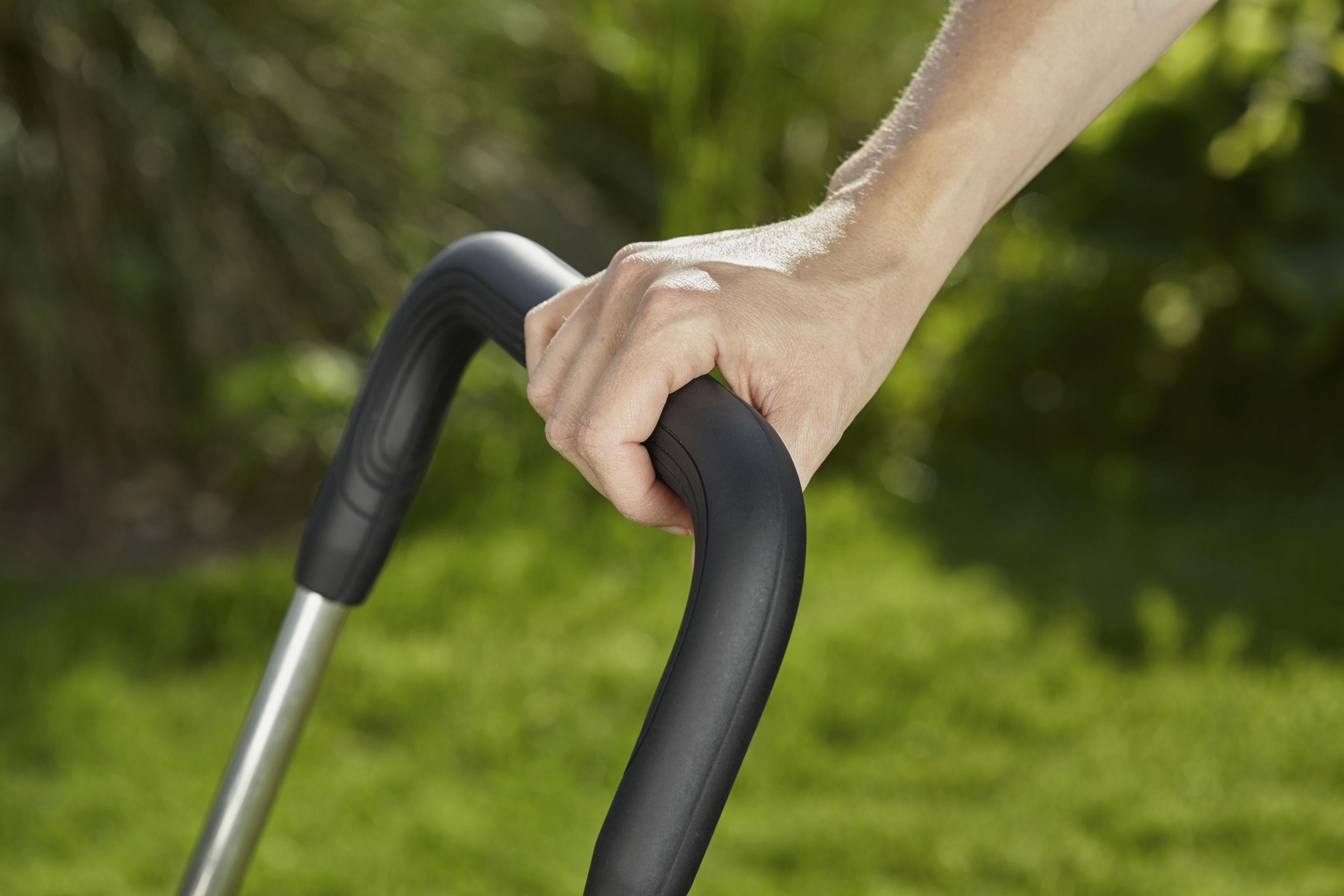 Eine Hand, die einen schwarzen, gebogenen Griff eines Rasenmähers vor einem grasbewachsenen Hintergrund hält.