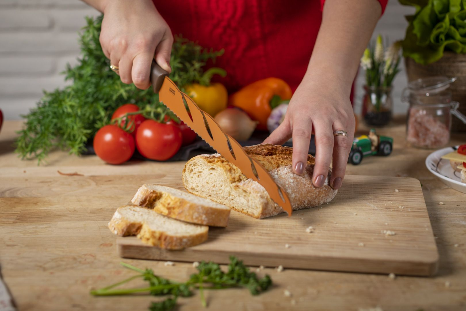 Osoba krojąca chleb na drewnianej desce pomarańczowym nożem, warzywa w tle.
