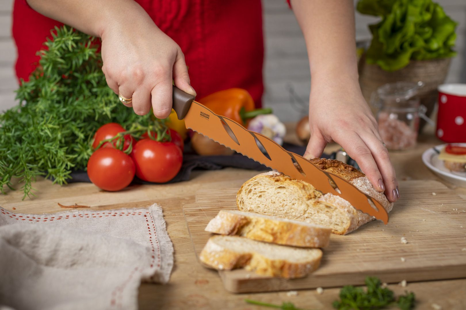 Osoba kroi chleb pomarańczowym nożem z ząbkami. Pomidory, zioła i warzywa na drewnianej desce.