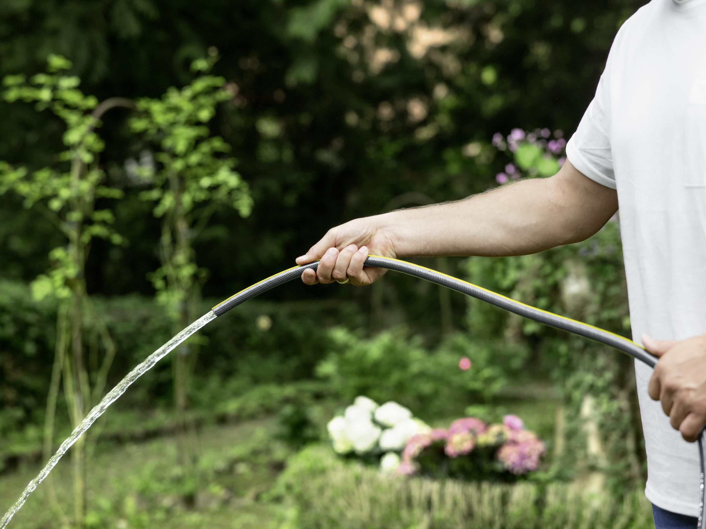 Osoba trzymająca wąż, woda tryska z węża w otoczeniu ogrodu.