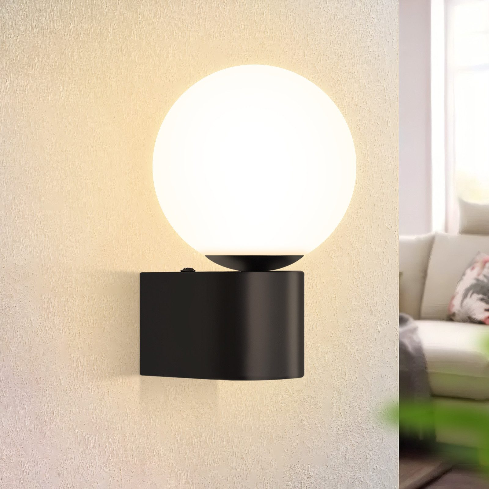 Una lampada da parete nera con luce bianca sferica. Il muro è beige.