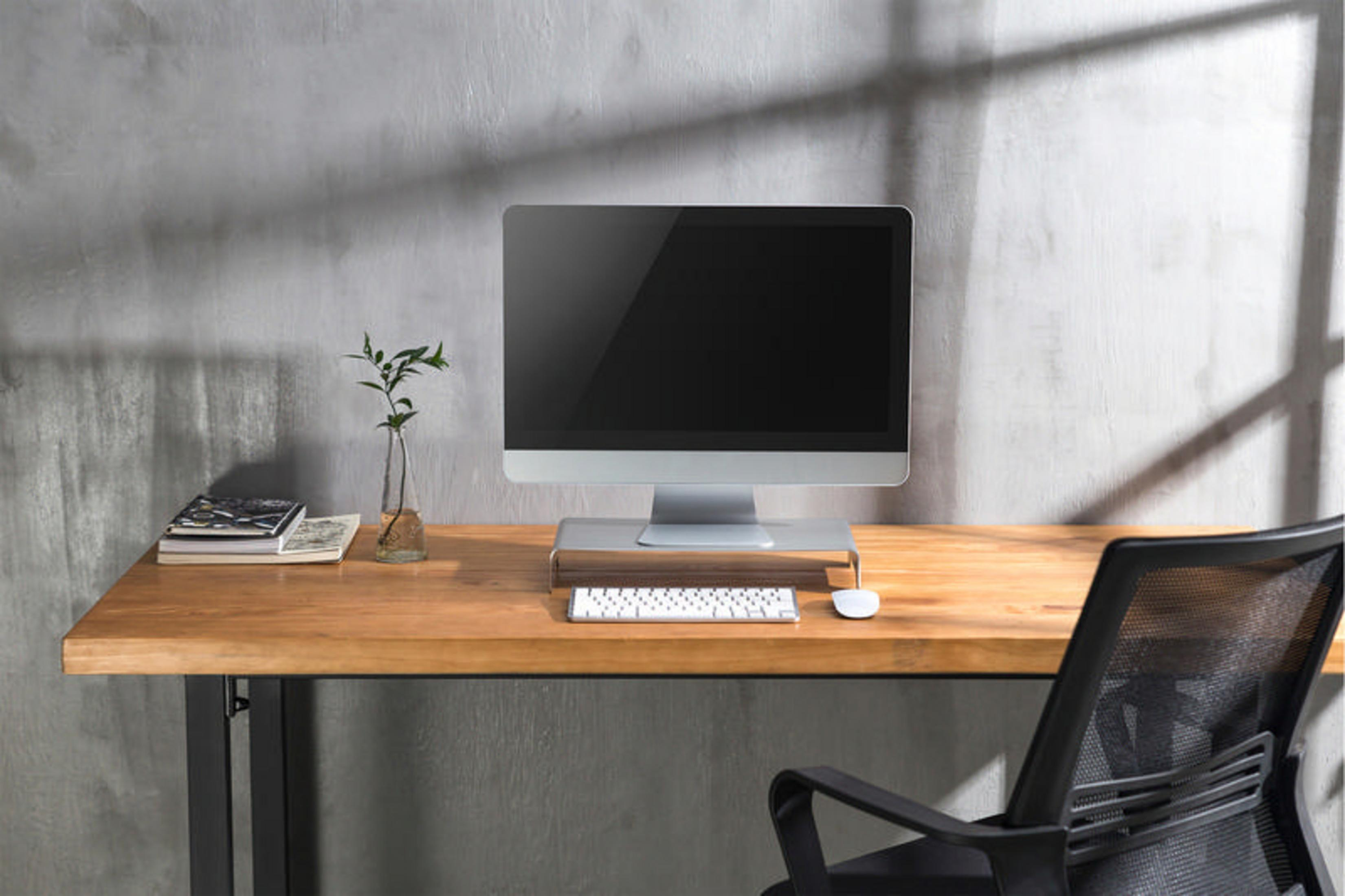 Biurko z monitorem komputera, klawiaturą i małą rośliną w szklanym wazonie. Szara ściana w tle.