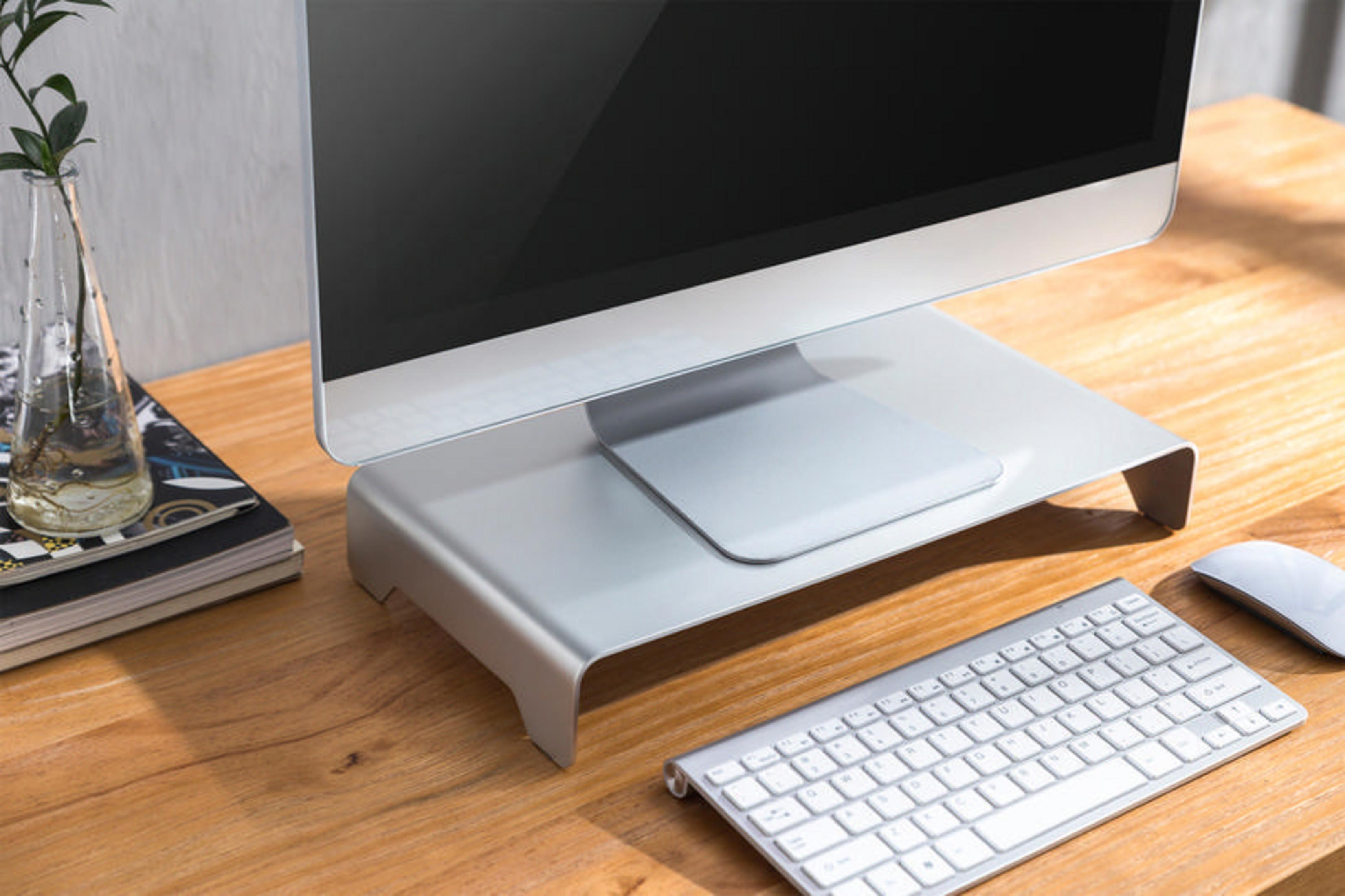 Monitor, klawiatura i mysz stoją na drewnianym biurku ze srebrną podstawką pod monitor. Po lewej wazon z zielonymi roślinami.