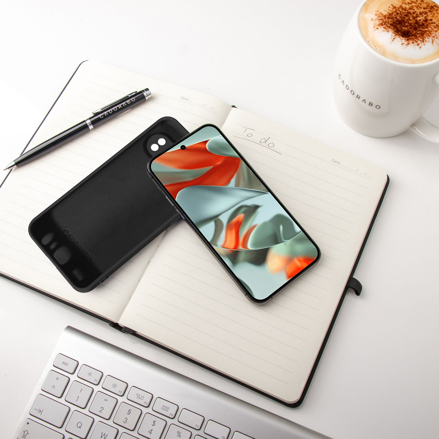 Notizbuch mit Telefon und Hülle, Stift und Kaffeetasse und eine Tastatur auf einem weißen Schreibtisch.