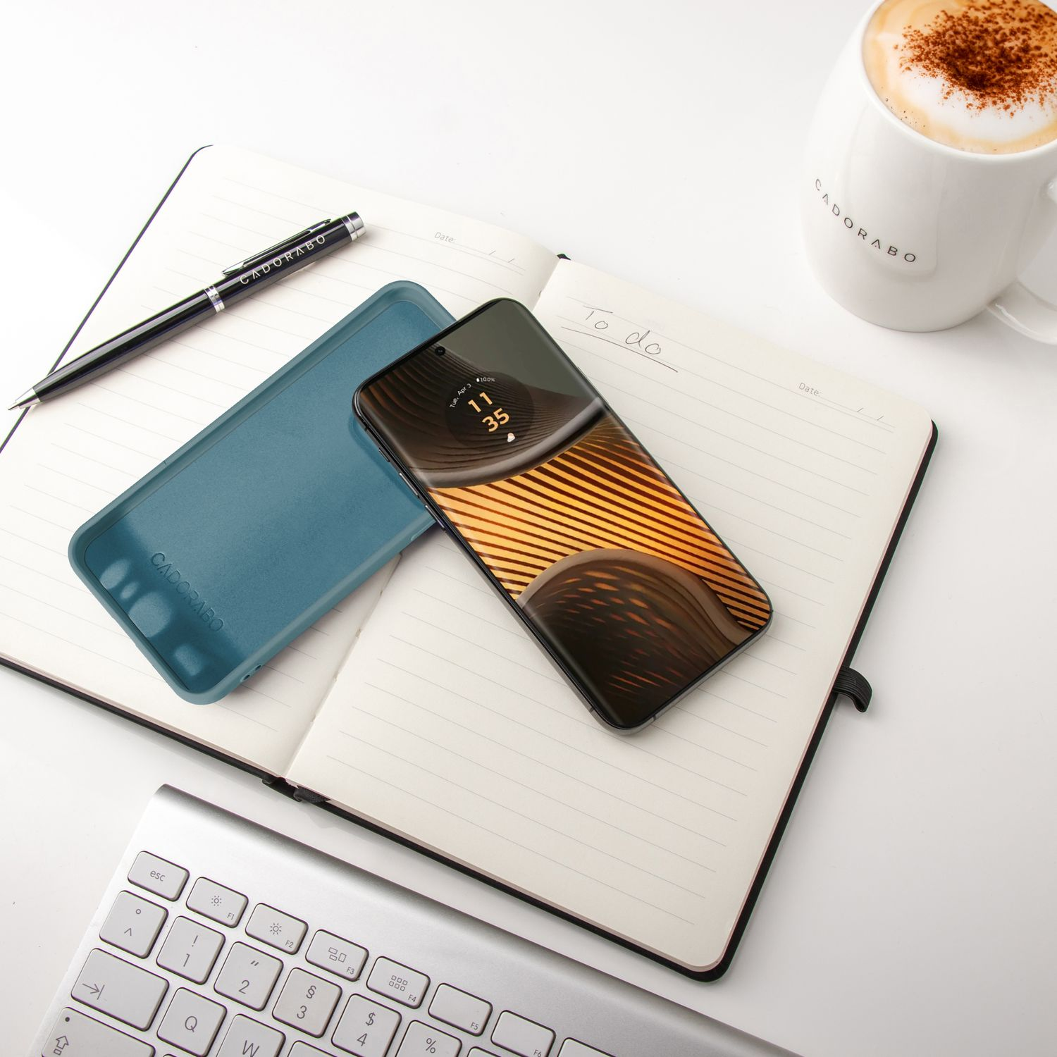 Telefon auf Notizbuch mit Hülle und Stift. Kaffeetasse und eine Tastatur. Weiße Oberfläche.