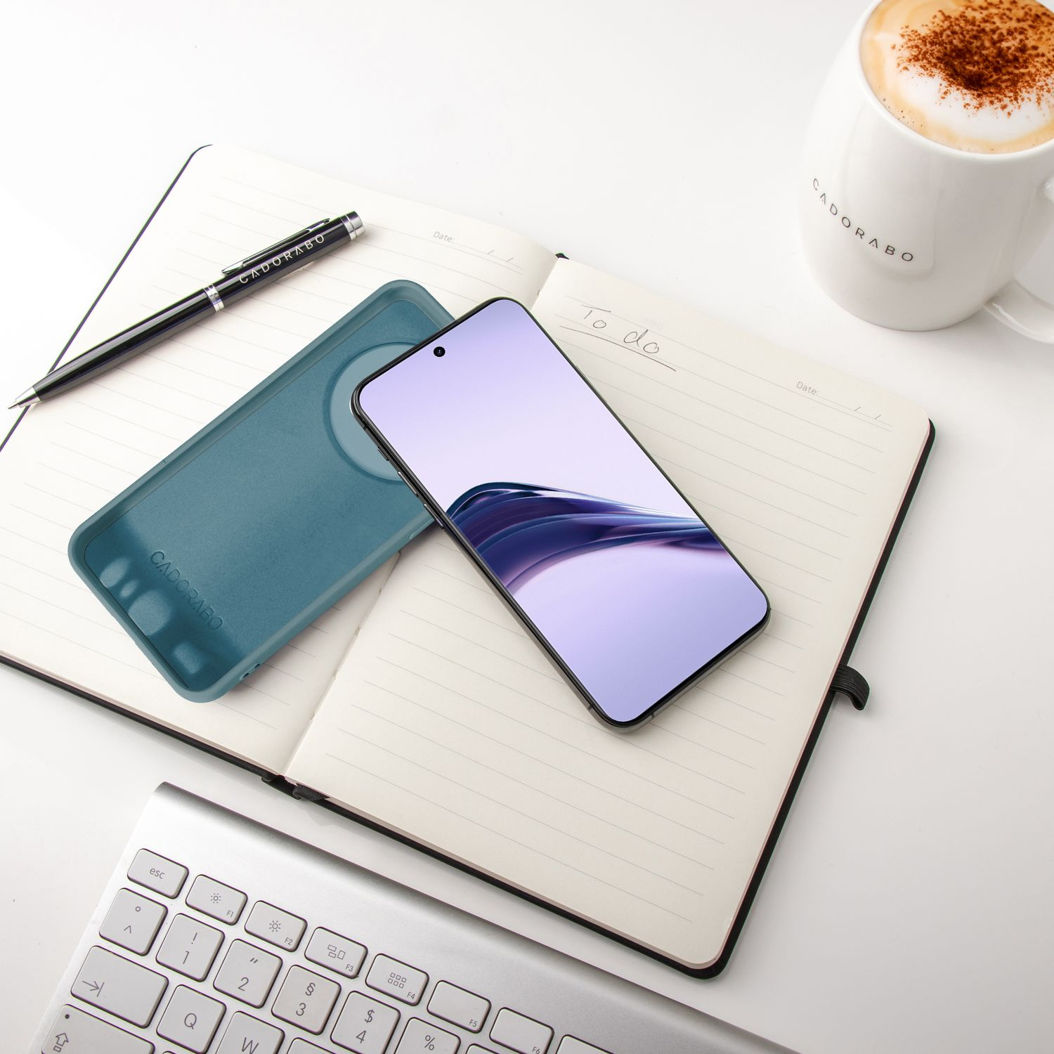 Notizbuch, Stift, Telefon, Handyhülle und eine Tasse Kaffee. Tastatur im Hintergrund. Weiße Oberfläche.