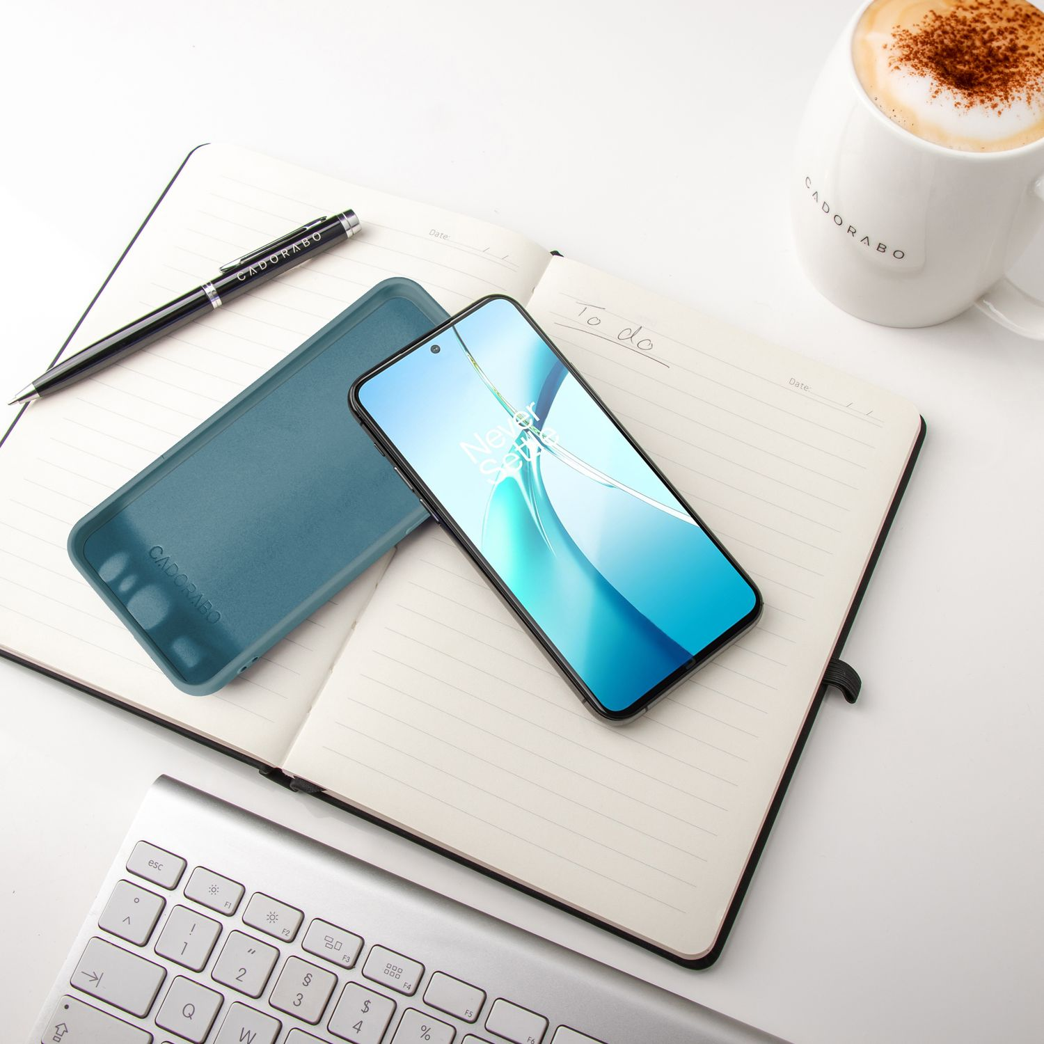 Notizbuch, Stift, Telefon, Handyhülle und eine Tasse Kaffee. Tastatur im Hintergrund. Weiße Oberfläche.
