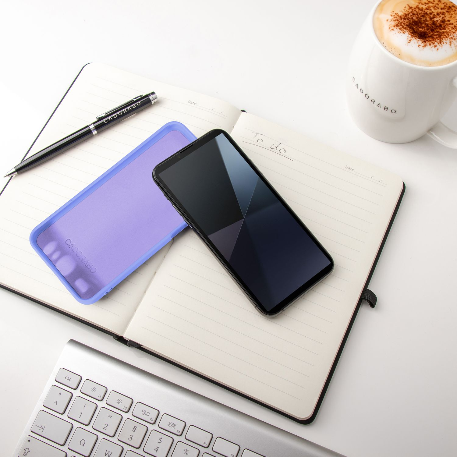 Ein Notizbuch, Telefon und Stift liegen auf einem weißen Schreibtisch. Eine Tasse Kaffee ist sichtbar. Die Handyhülle ist ebenfalls vorhanden.