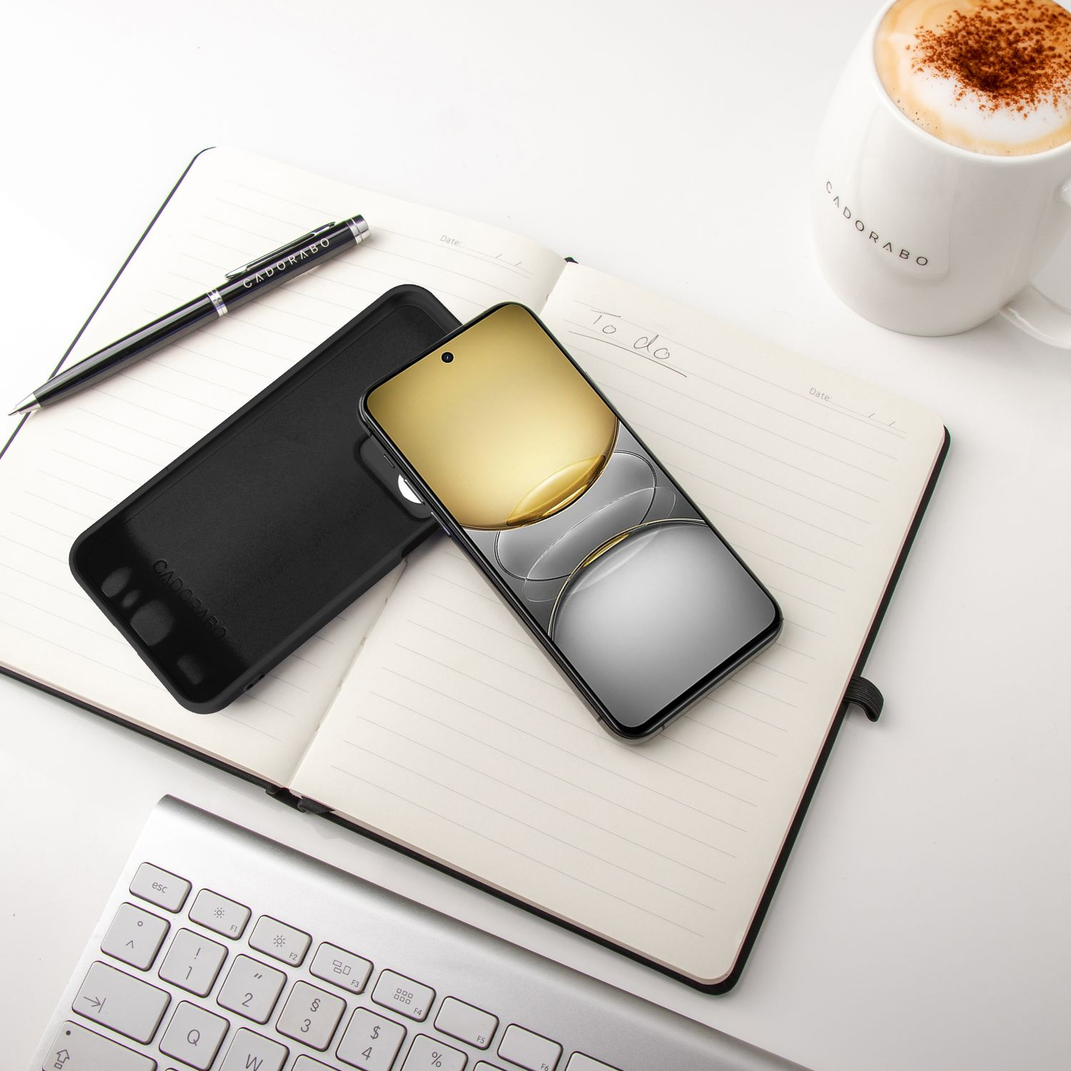 Telefon in schwarzer Hülle auf offenem Notizbuch mit Stift und Kaffee. Weiße Tastatur im Hintergrund.