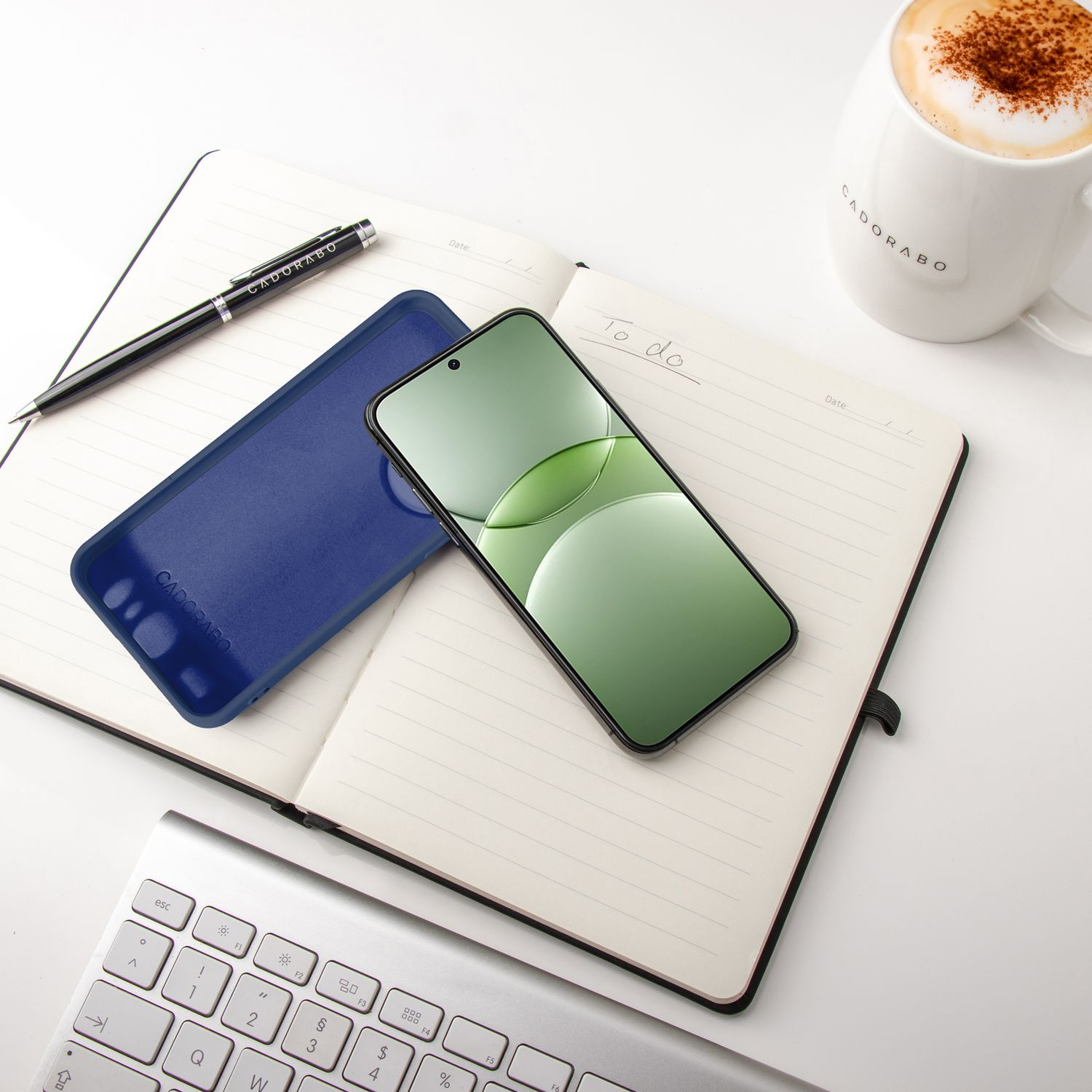 Notizbuch mit Telefon & blauer Hülle. Stift und Becher stehen auf dem Tisch. Tastatur in der Ecke.