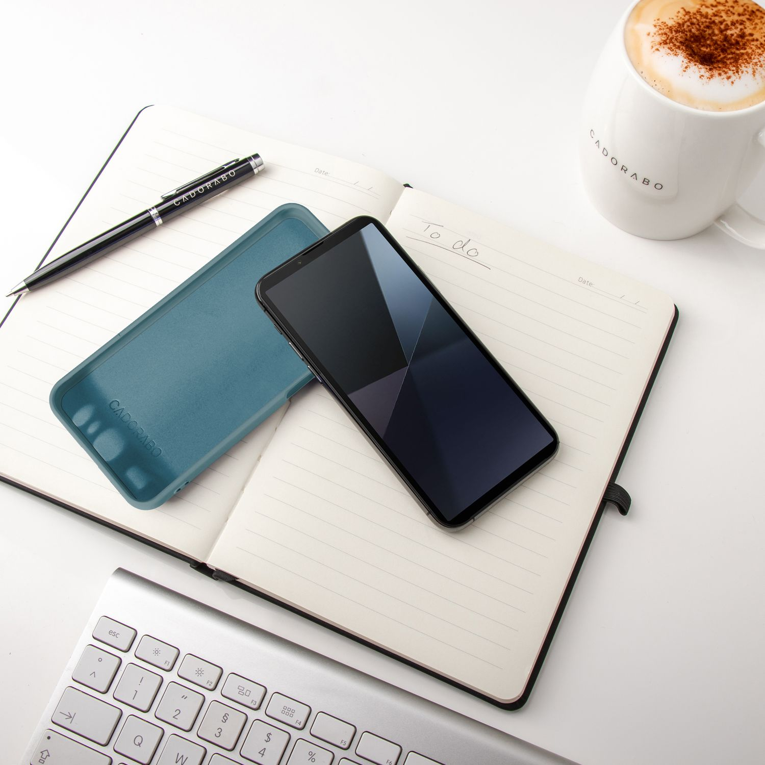 Notizbuch mit Telefon, Stift und Tasse. Das Telefon und die Hülle liegen neben einer Kaffeetasse.