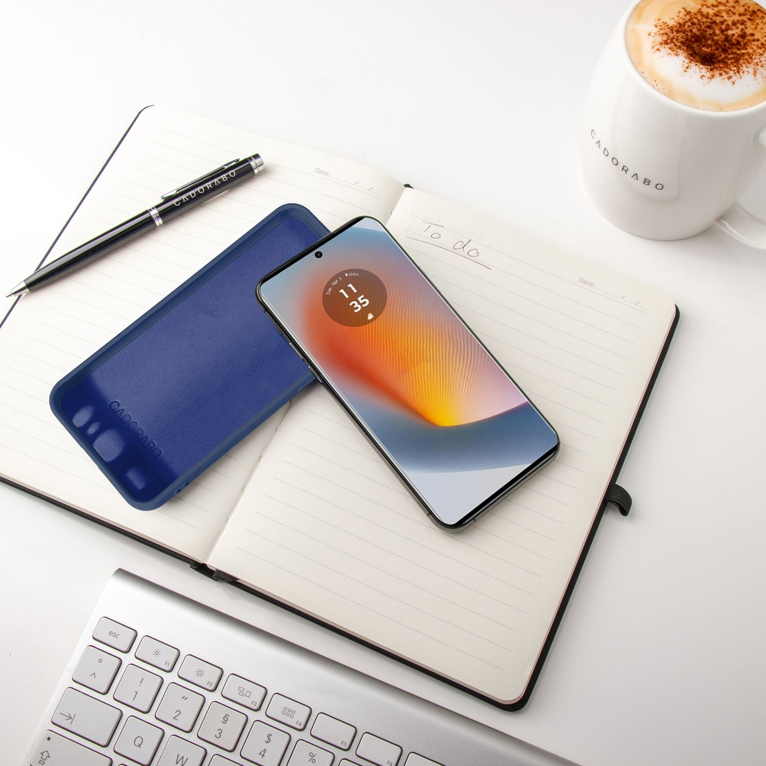 Telefon in Hülle auf Notizblock, neben Stift und Kaffeetasse. Weißes Schreibtischsetting. Telefon zeigt Datum und Uhrzeit.