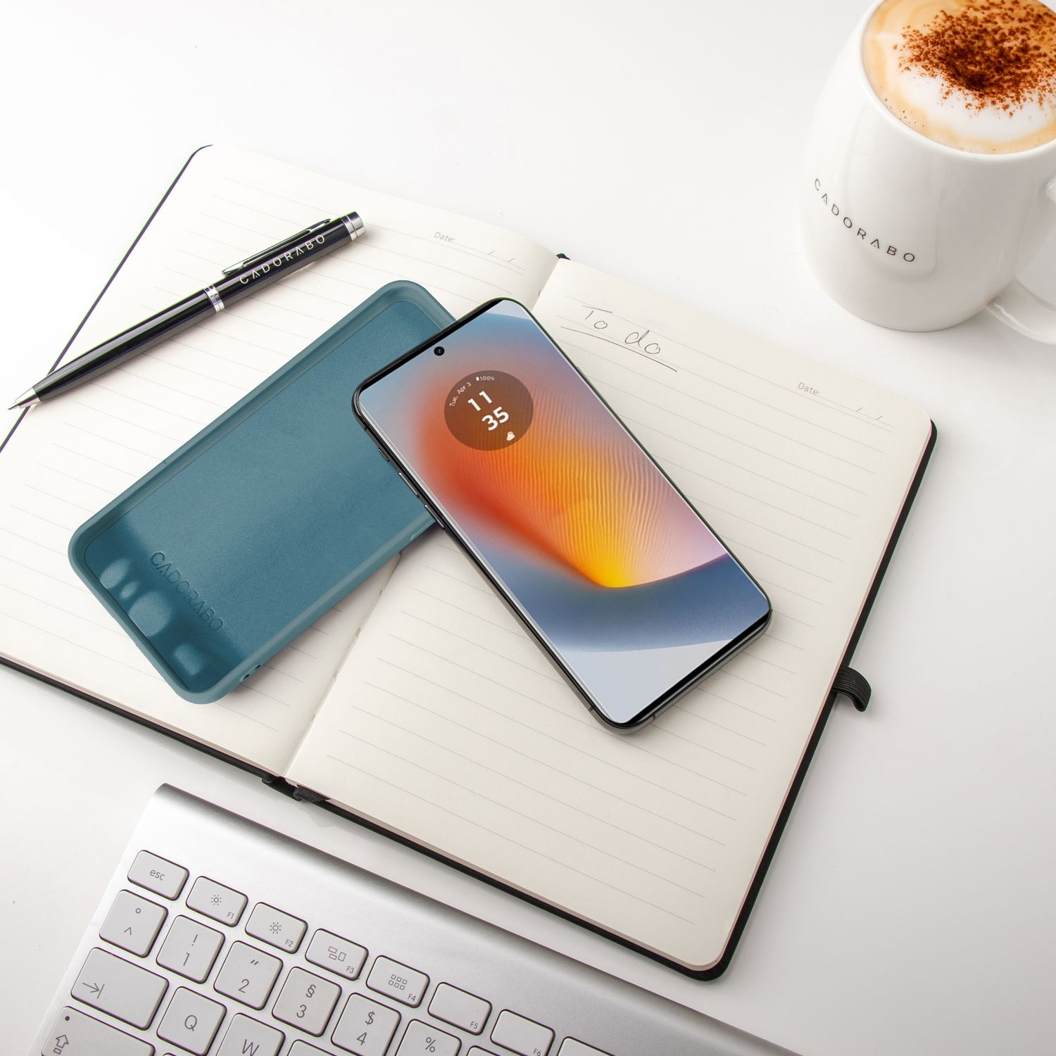 Schreibtischszene, Telefon zeigt Zeit, Notizblock, Stift und eine Tasse Kaffee auf weißer Oberfläche.