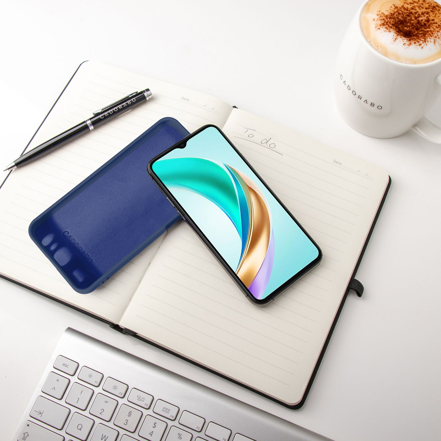 Notizbuch mit Smartphone und blauer Handyhülle, Stift und Kaffeetasse auf weißem Tisch.