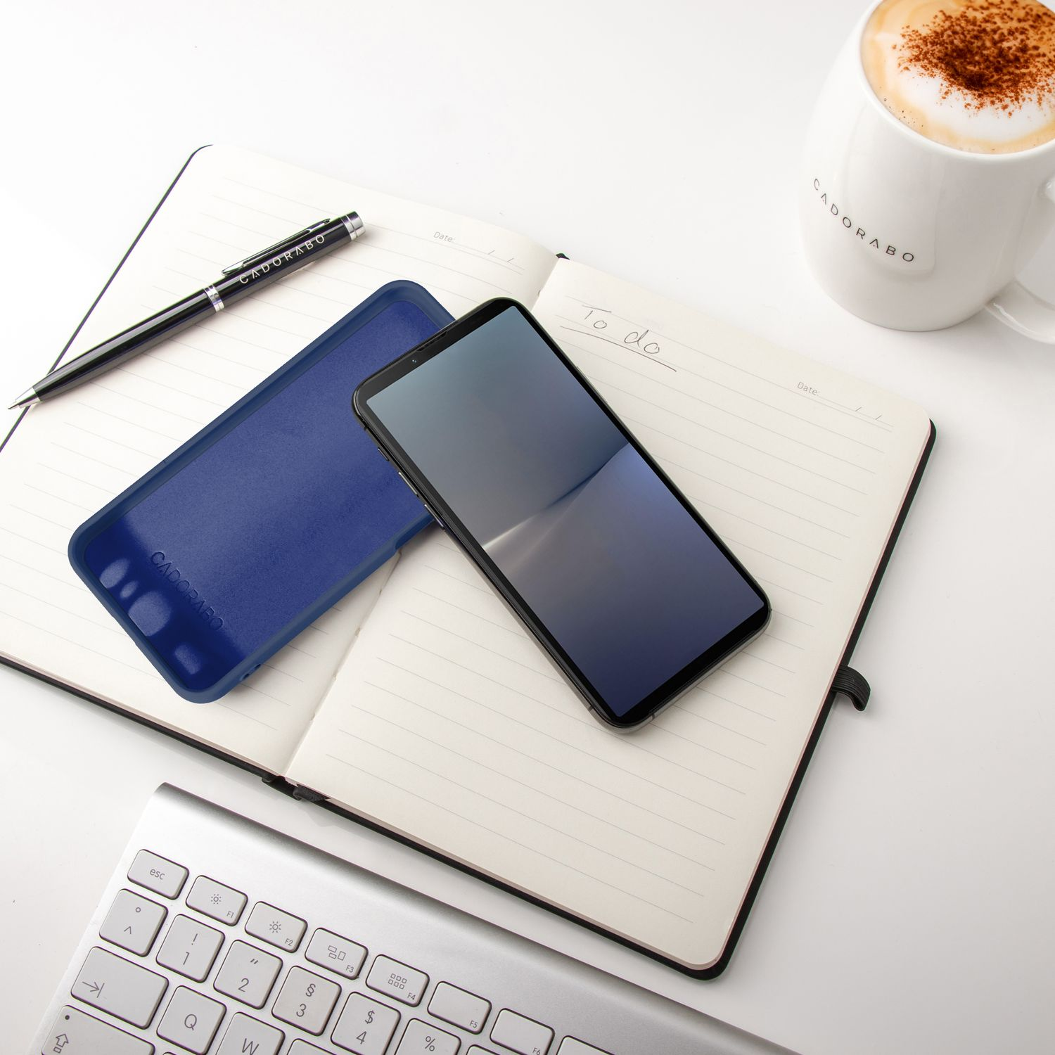 Ein Telefon und eine Handyhülle auf einem offenen Notizbuch, mit einem Stift und einer Tasse Kaffee.
