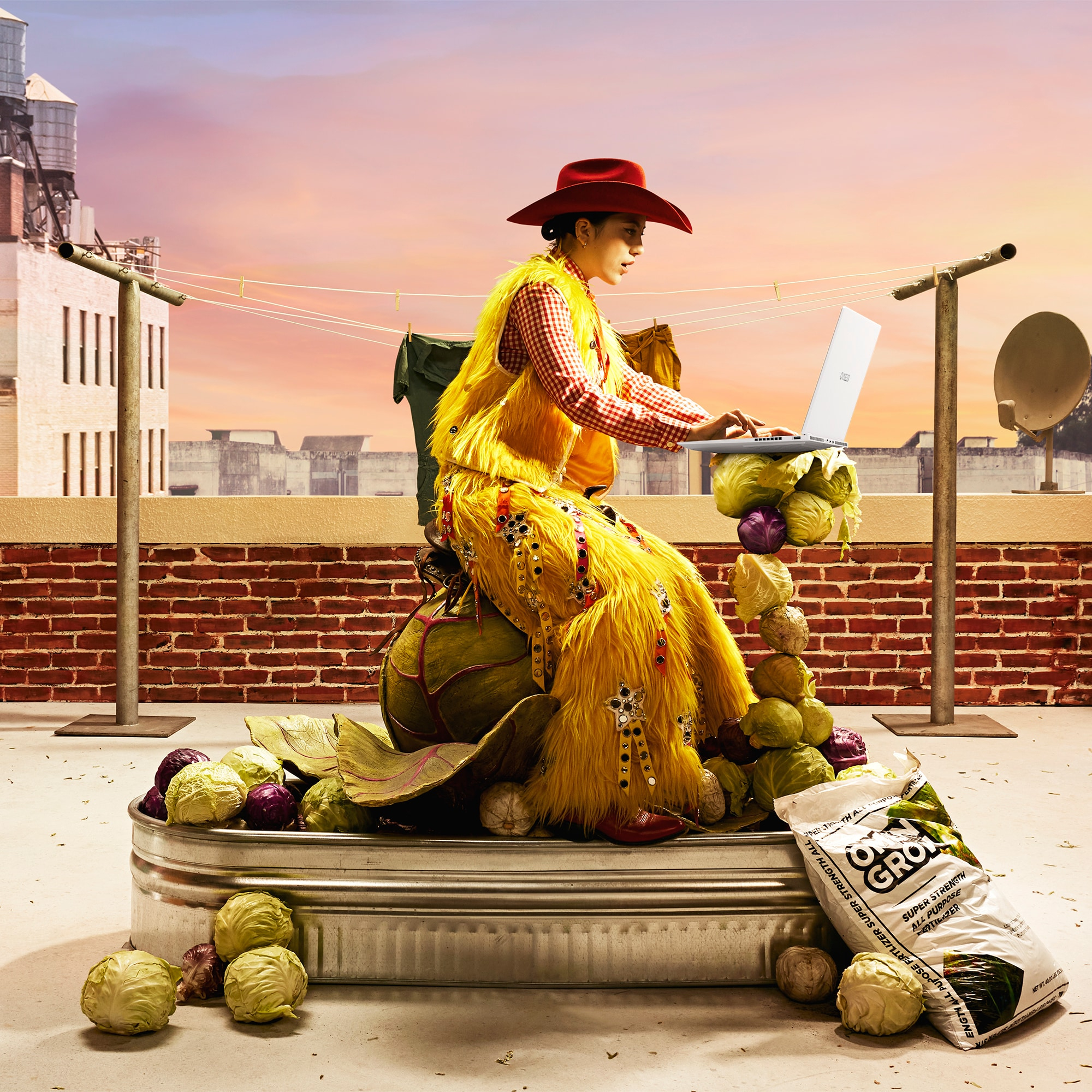 Frau in gelbem Outfit benutzt Laptop auf Dach mit Kohl und Backsteinmauer.