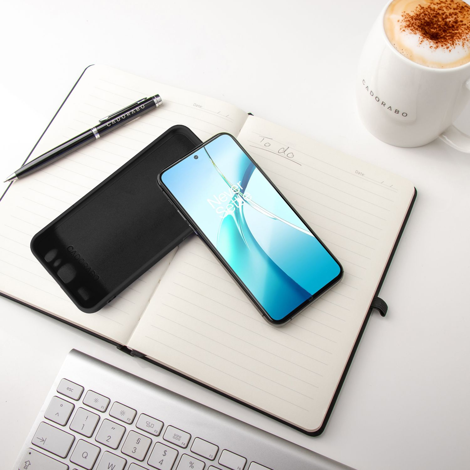 Notizbuch mit Telefon, Hülle, Stift und Kaffeetasse auf weißem Tisch.
