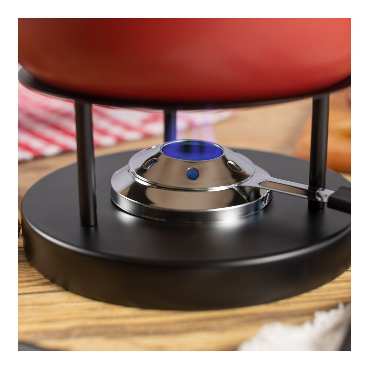Ein Fondue-Set mit einem roten Topf. Die Basis ist schwarz mit einem metallischen Brenner. Es steht auf einem Holztisch.