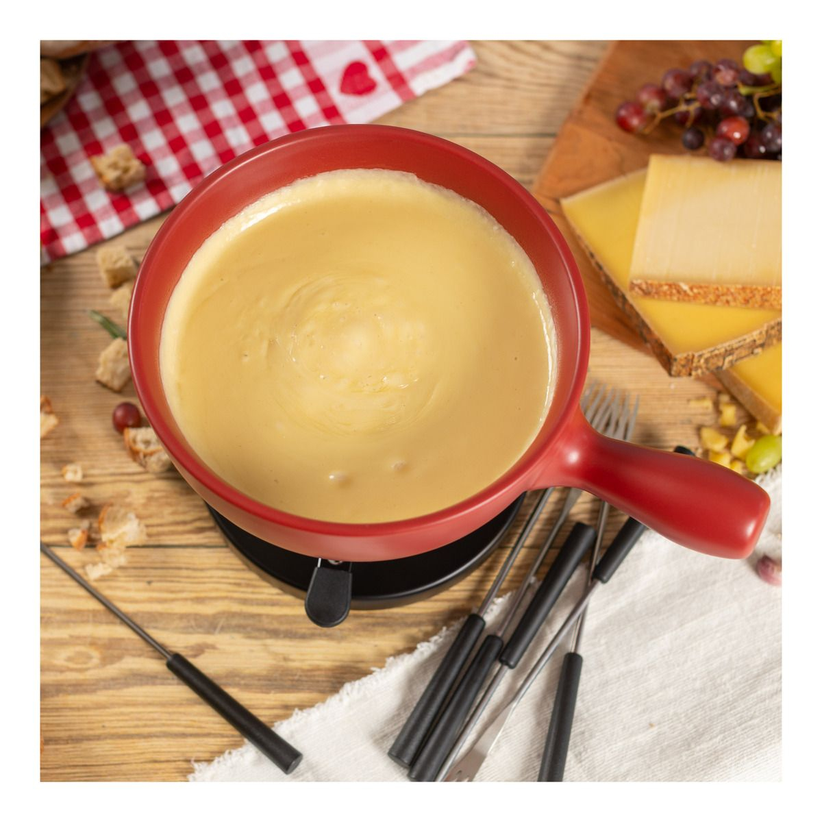 Fondue-Topf mit geschmolzenem Käse und Gabeln, auf Holztisch, rot-weißes Tuch.