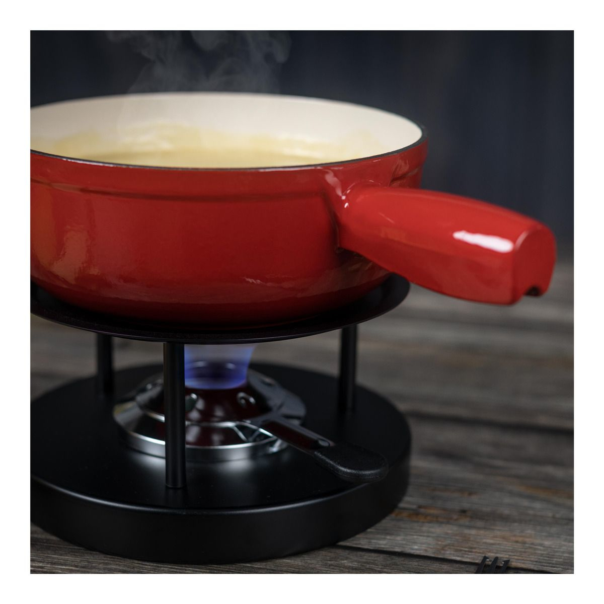 Roter Fonduetopf mit Dampf, auf schwarzem Ständer mit blauer Flamme, auf Holzoberfläche.