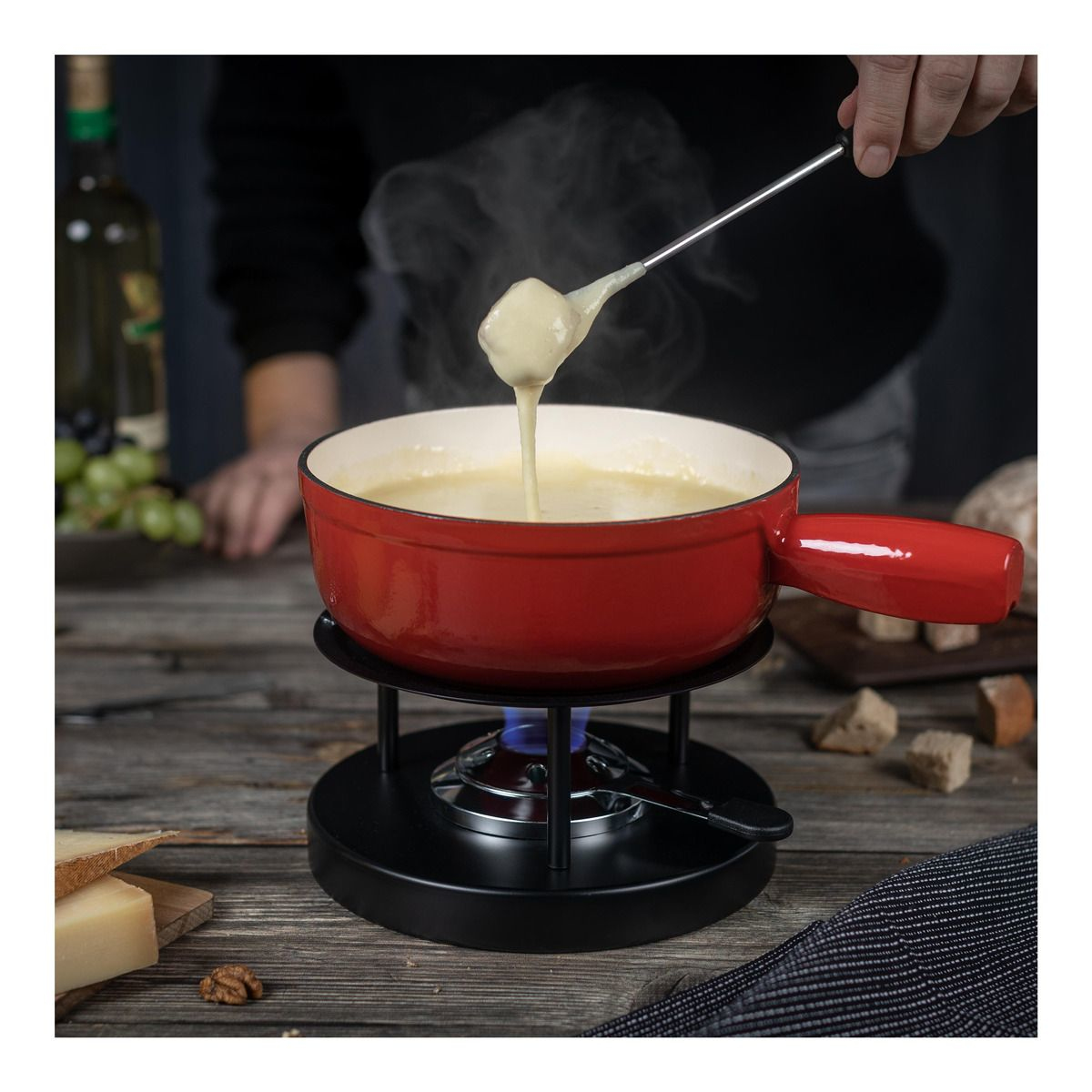 Roter Fonduetopf auf einem Brenner, Käse wird eingetaucht. Holztisch und Zutaten im Hintergrund.