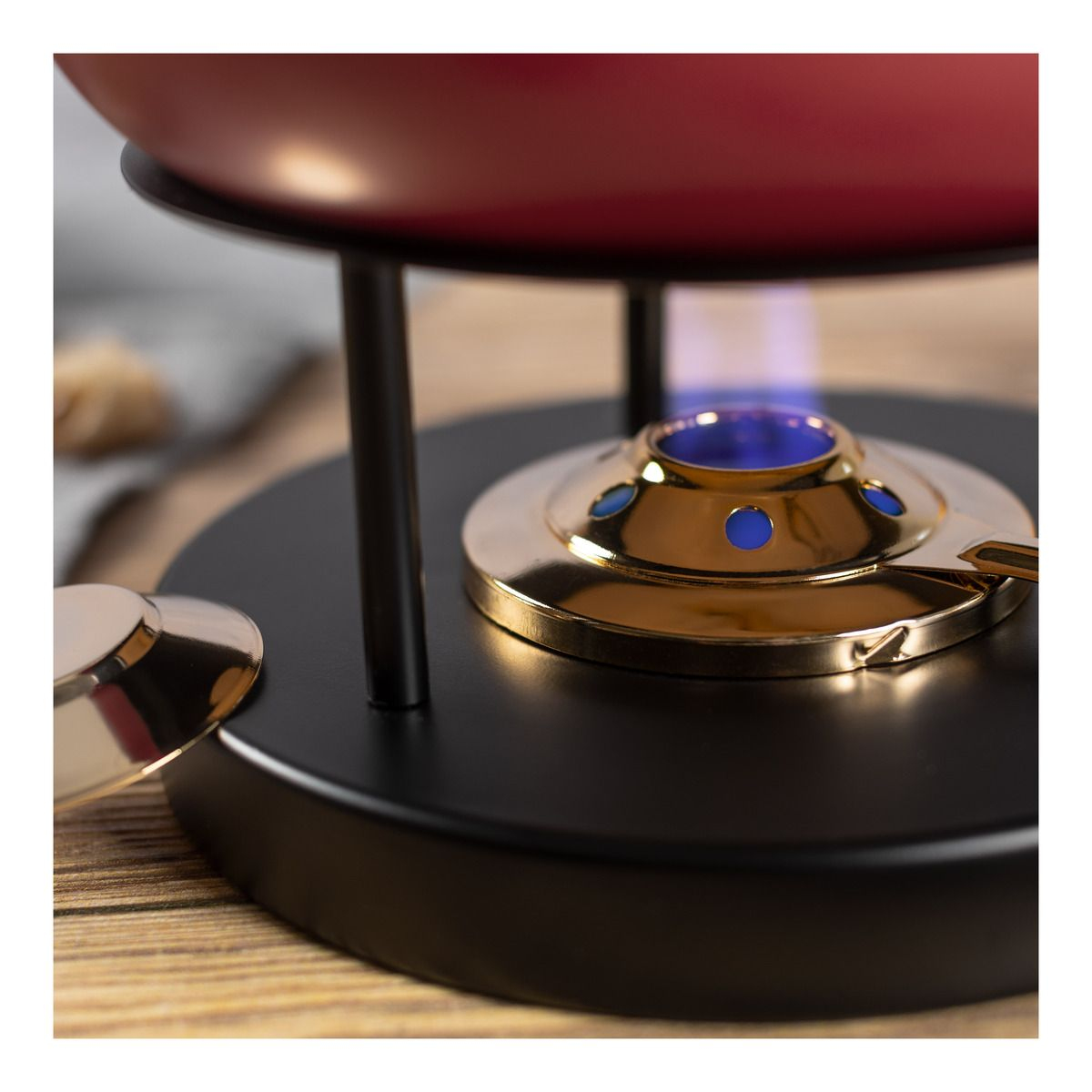 Roter Fonduetopf auf schwarzem Ständer mit blauer Flamme und goldenem Brenner auf einem Holztisch.