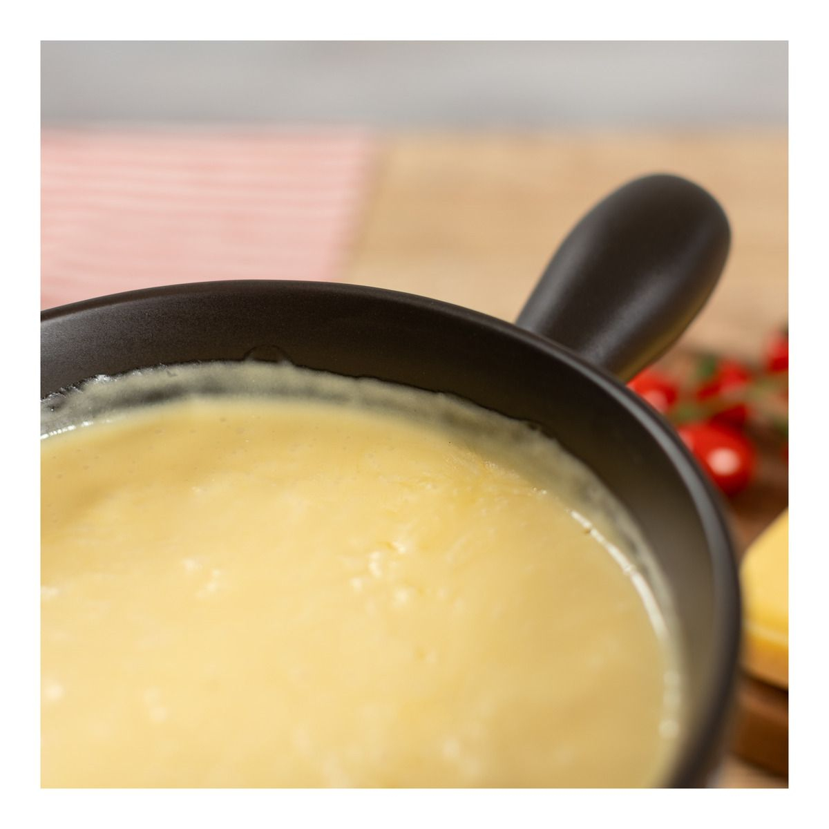 Nahaufnahme eines Fonduetopfes mit geschmolzenem Käse. Holztisch und Tomaten im Hintergrund.
