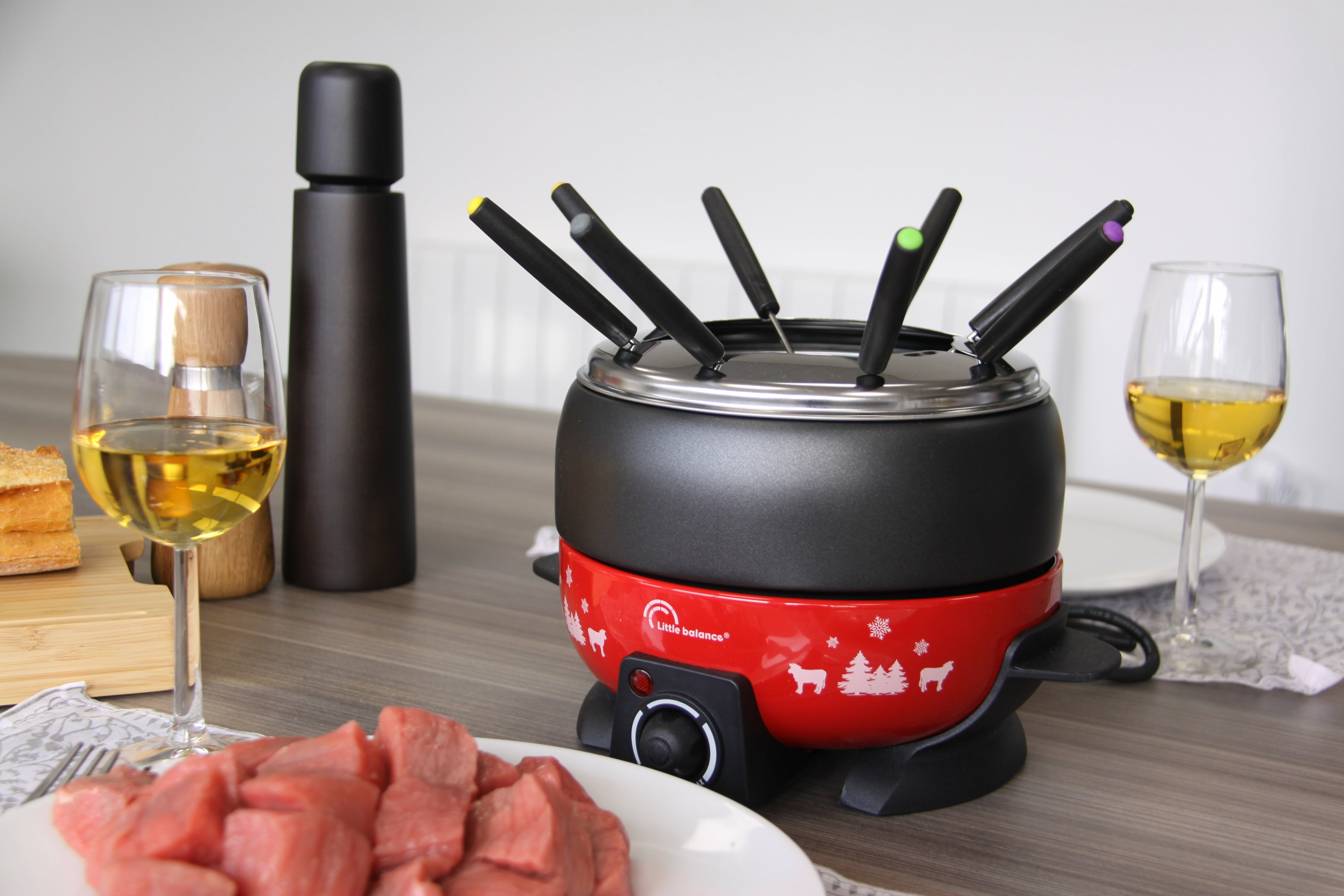 Fondue-Set mit Gabeln, Weingläsern und Fleischwürfeln auf einem Holztisch.