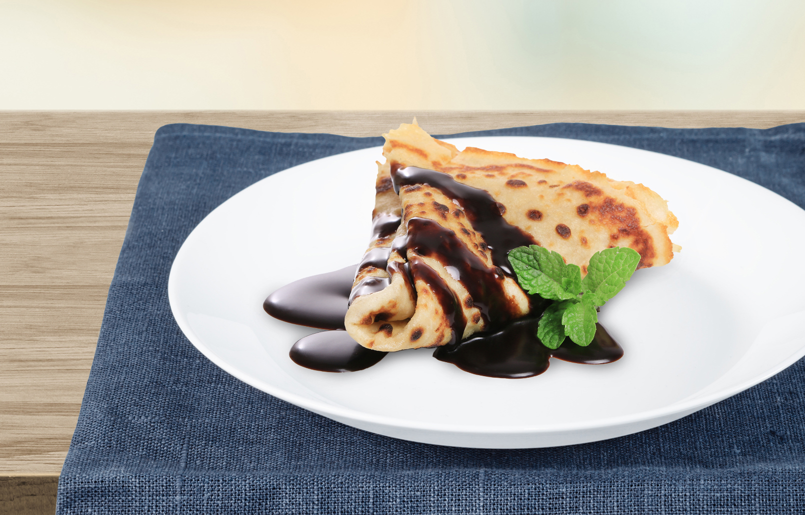 Crêpe mit Schokoladensauce und Minze auf einem weißen Teller, auf einer blauen Serviette.