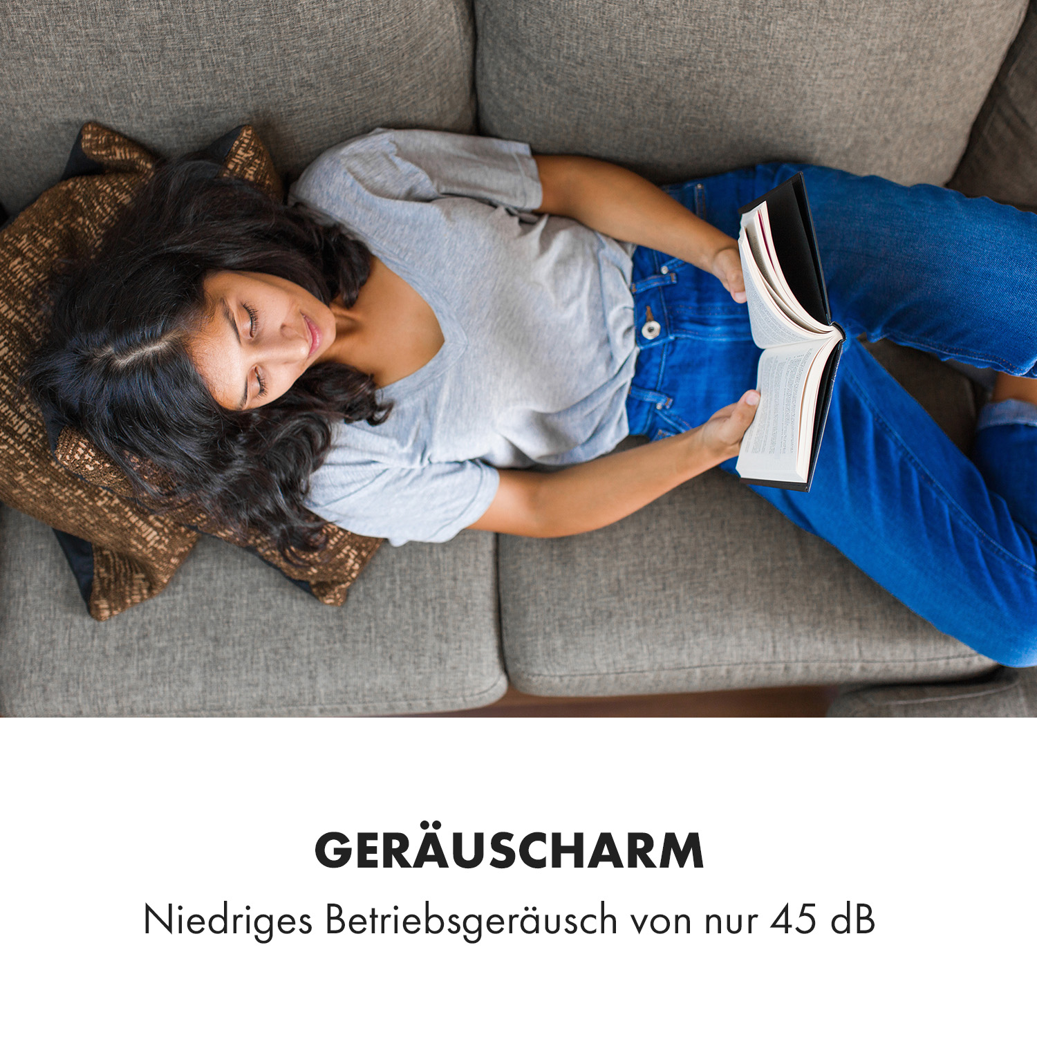 Frau liegt auf Sofa und liest Buch. Graues Sofa, braunes Kissen, blaue Jeans. Lächelt, entspannt.