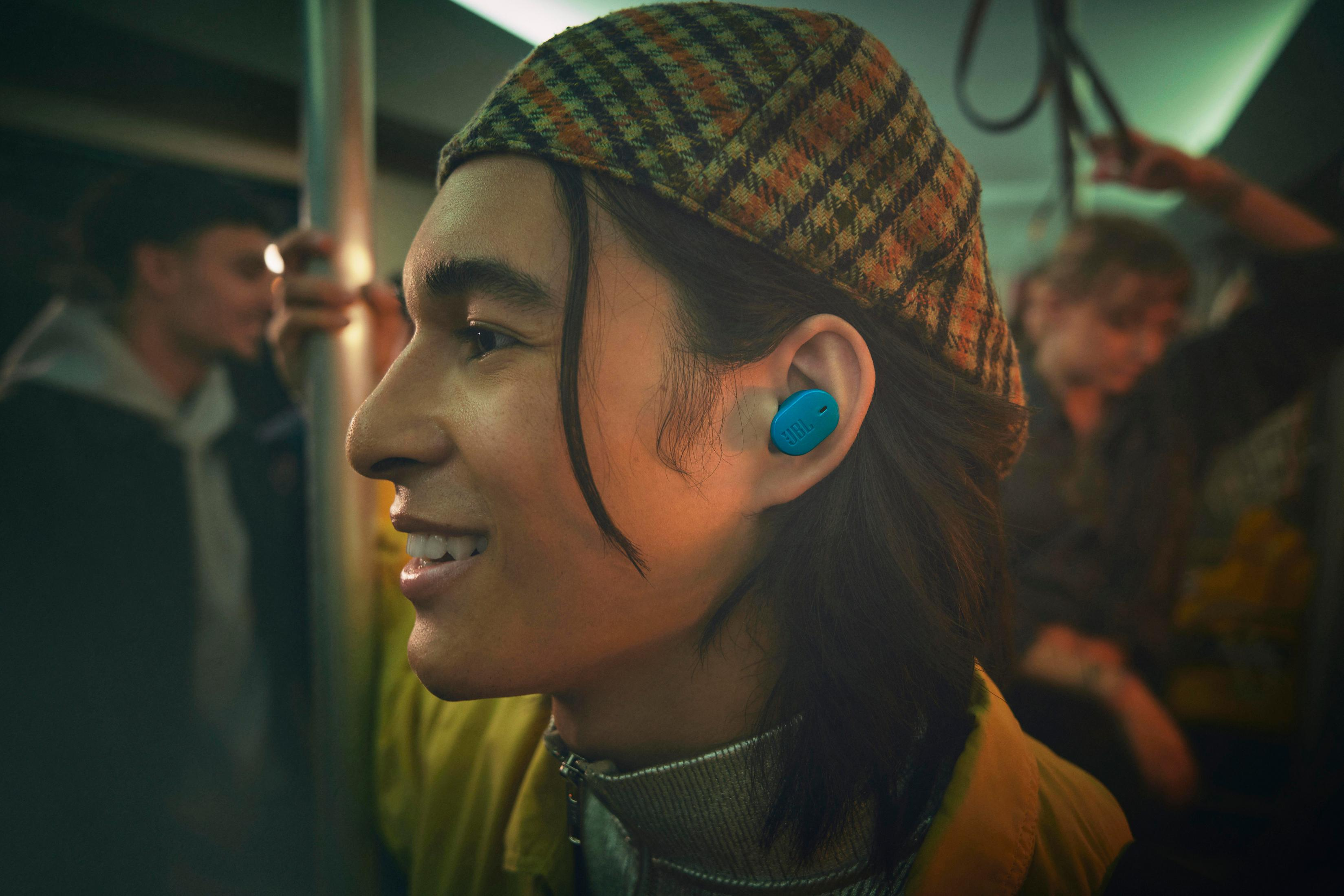 Un giovane con auricolari blu in metropolitana. Sorride e indossa un cappello.