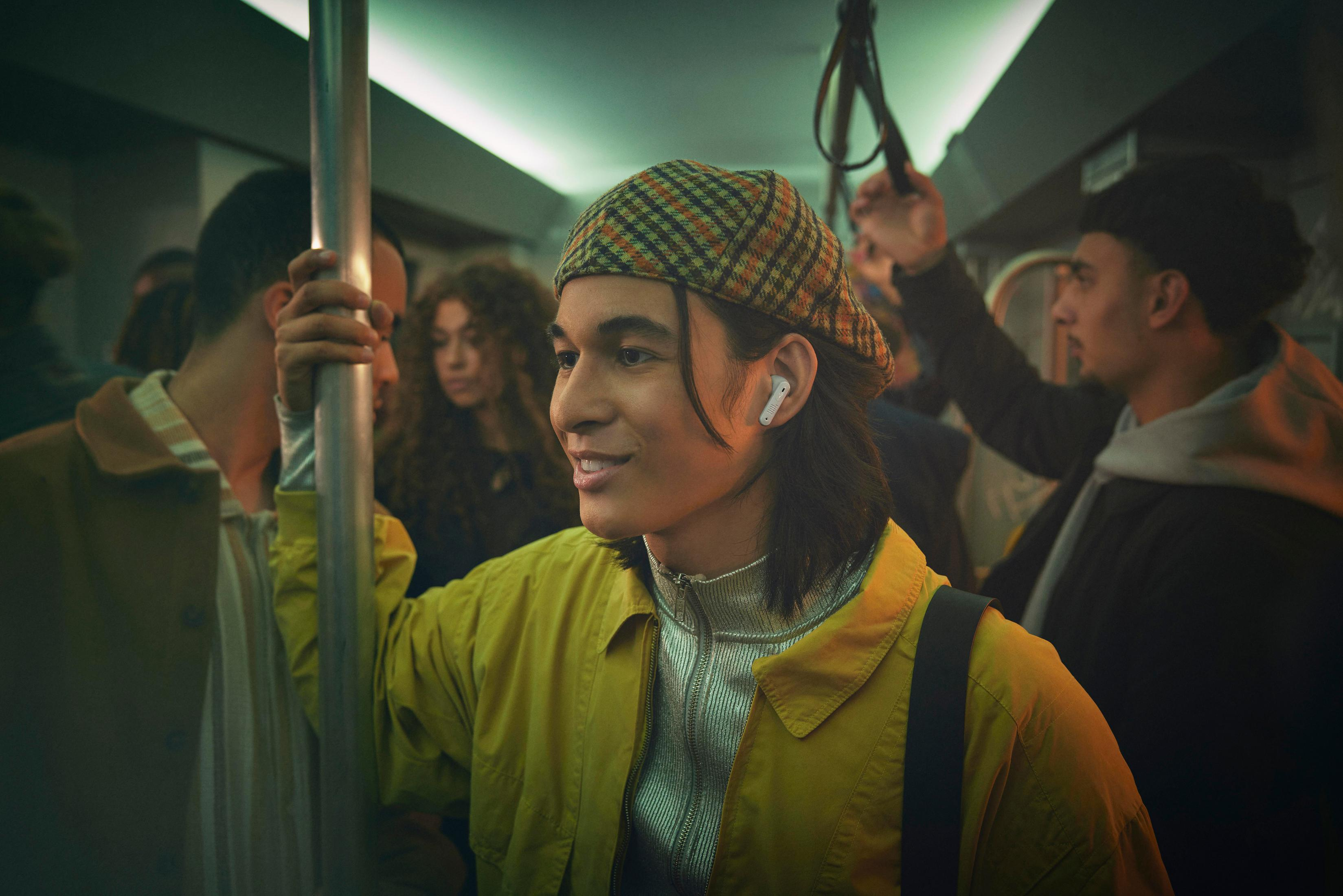 Giovane con un cappello su un treno, che indossa auricolari. Circondato da altri passeggeri.