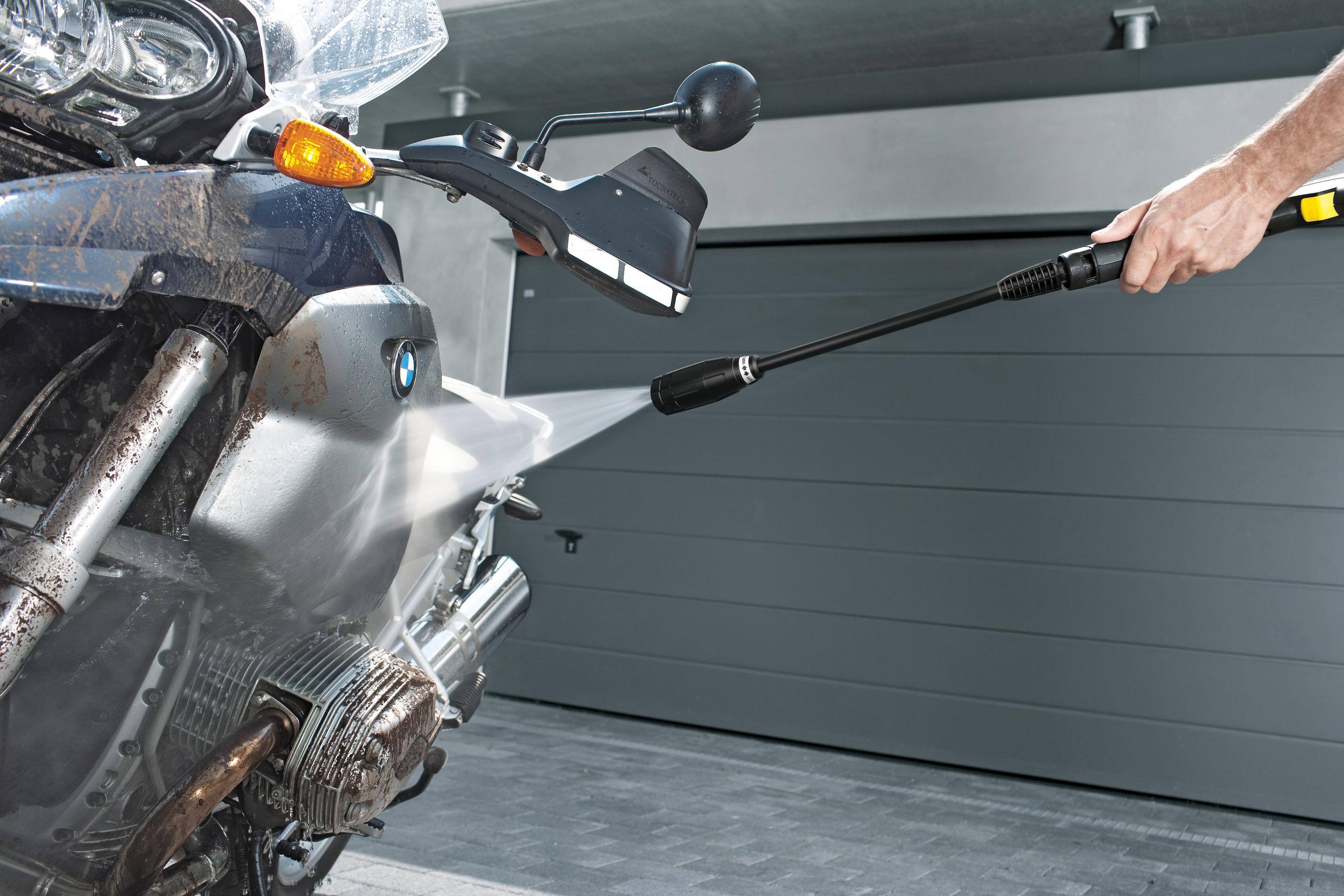 Een motorfiets wordt gewassen met een hogedrukreiniger. Een man houdt de wasser vast. De achtergrond is een garage.