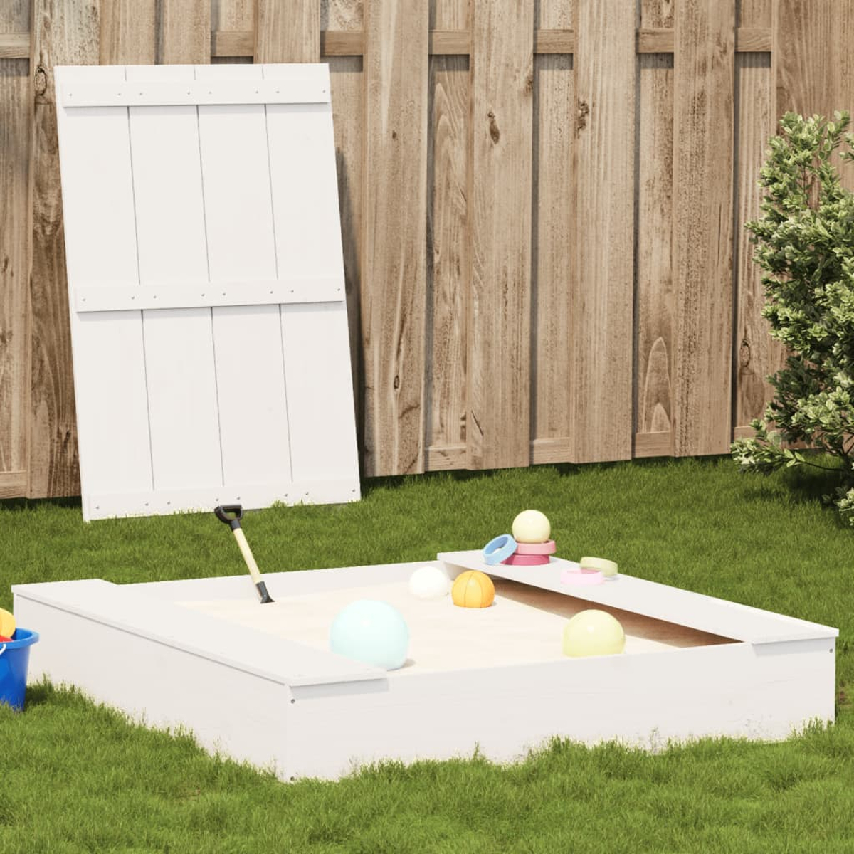 Sabbiera in legno bianco con coperchio, giocattoli sparsi sulla sabbia, erba verde.