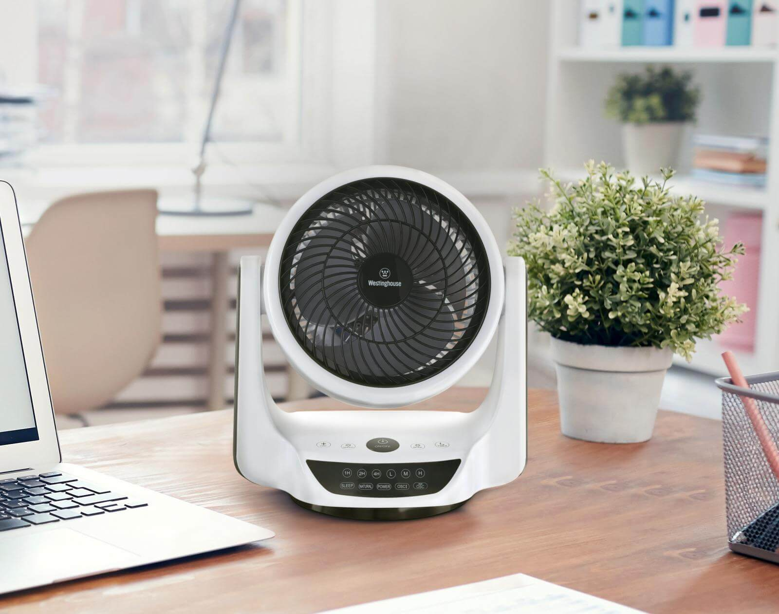 Ein weiß-schwarzer Ventilator auf einem Holzschreibtisch mit Laptop und grüner Pflanze im Hintergrund.