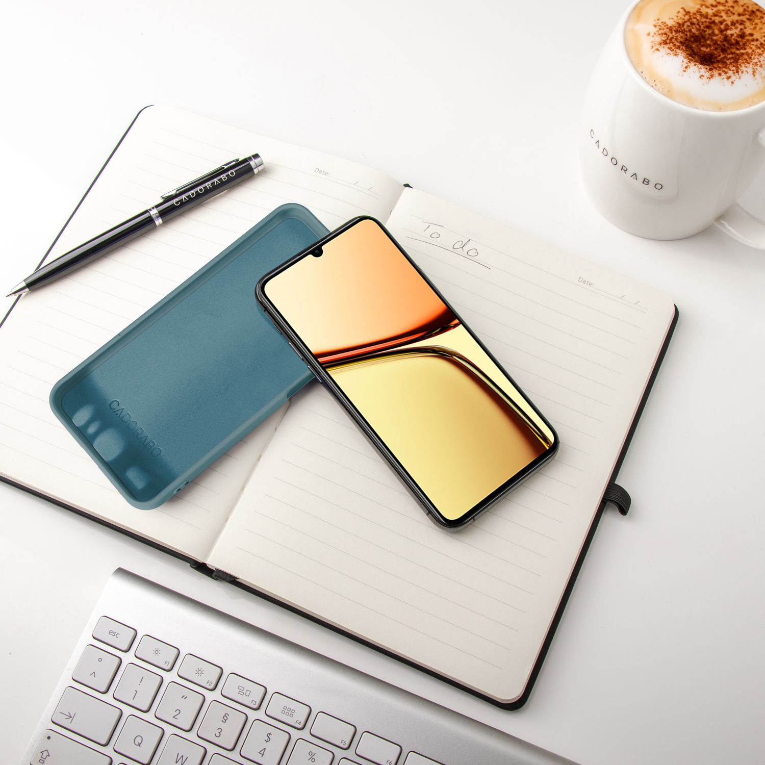 Ein Telefon mit türkisfarbener Hülle, ein Stift, ein Notizblock mit Schrift und eine Tasse auf einem weißen Schreibtisch.