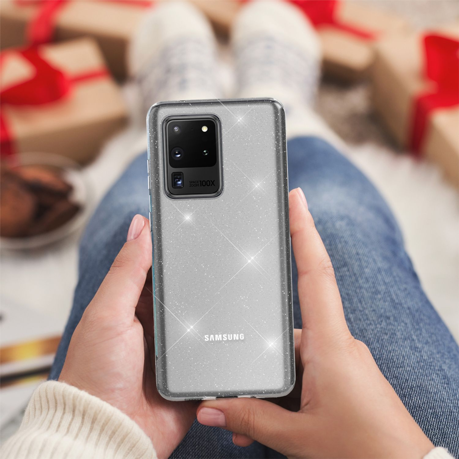 Hände halten ein Telefon in einer Glitzerhülle, sitzen auf einem Schoß mit Geschenken im Hintergrund.