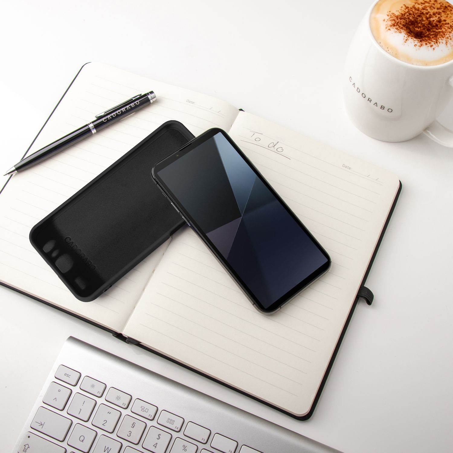 Telefon, Hülle, Stift und Kaffee auf einem Notizbuch. Tastatur im Vordergrund. Weißer Hintergrund, Draufsicht.
