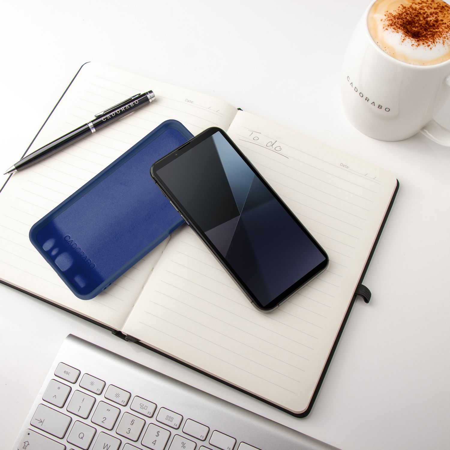 Telefon in Hülle auf offenem Notizbuch. Stift, Handyhülle und Kaffeetasse auf weißer Oberfläche.