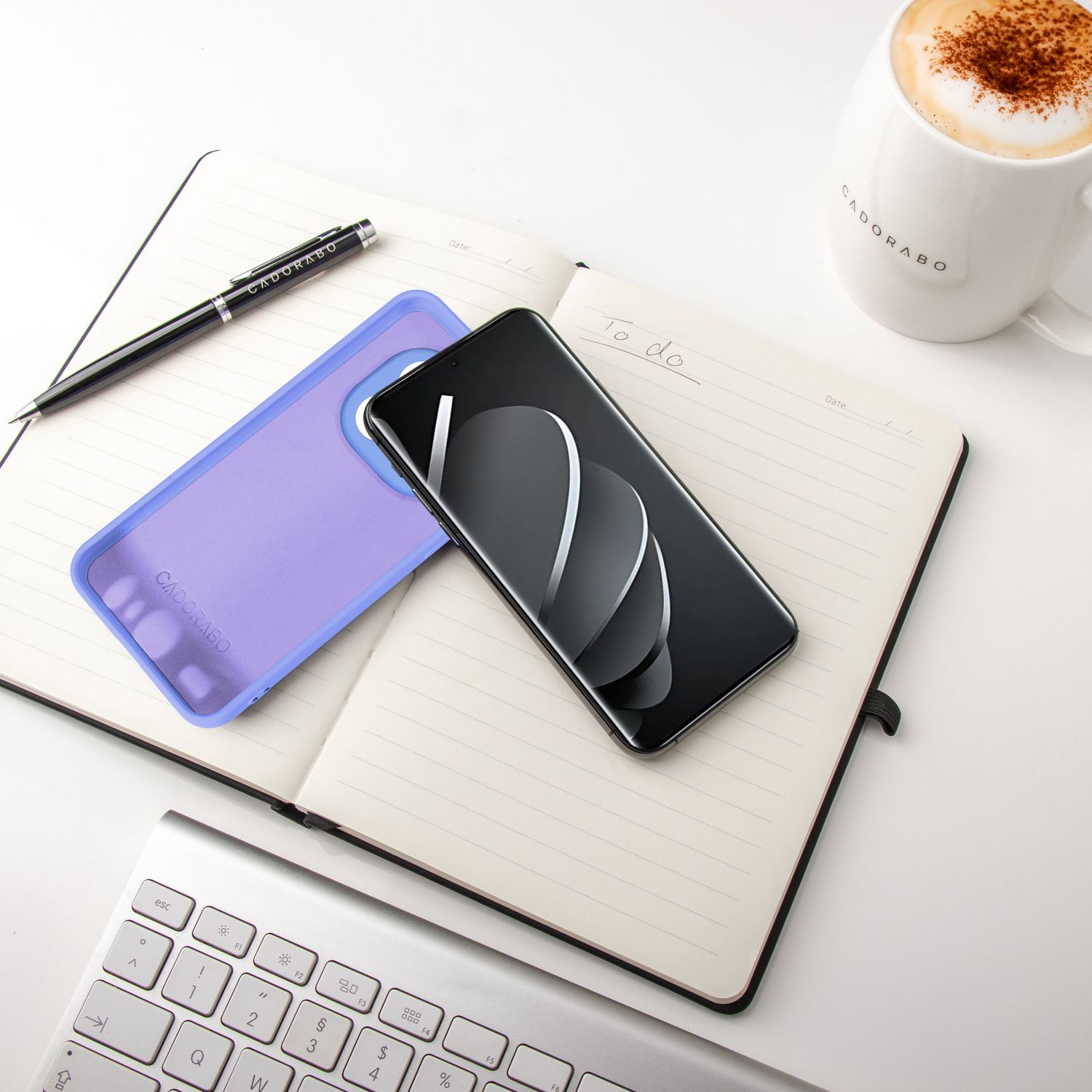 Ein Telefon und eine Hülle auf einem Notizbuch. Ein Stift und ein Becher sind vorhanden. Die Tastatur ist im Hintergrund.