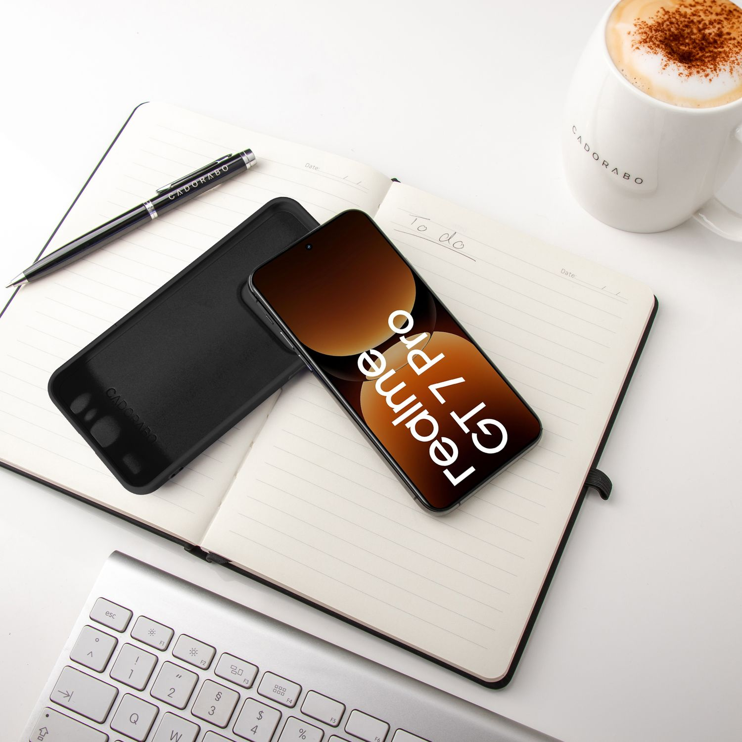 Ein Telefon mit schwarzer Hülle und eine Handyhülle liegen auf einem Notizbuch. Ein Stift und eine Tasse sind ebenfalls auf dem Tisch.