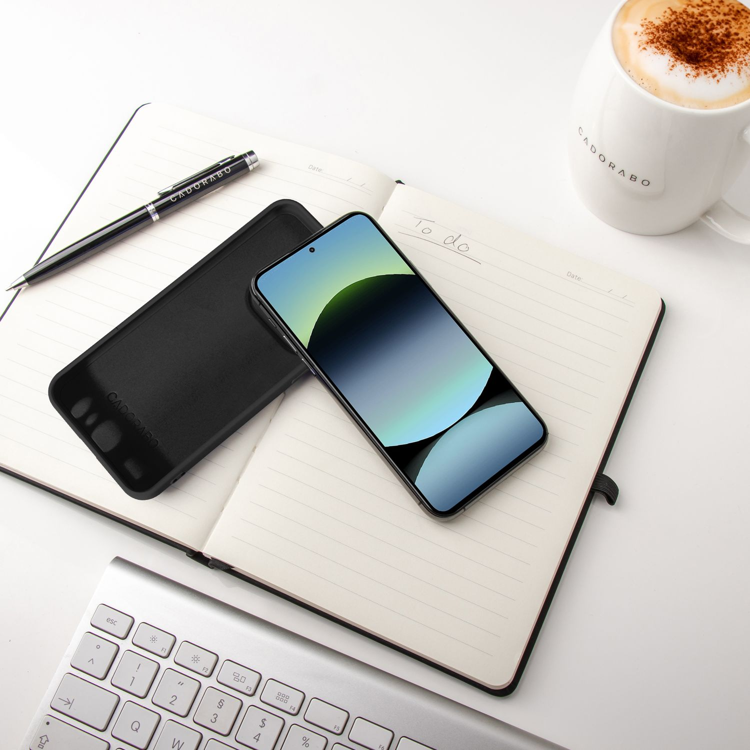 Offenes Notizbuch, Telefon mit buntem Bildschirm, Stift und Kaffeetasse auf weißer Oberfläche.