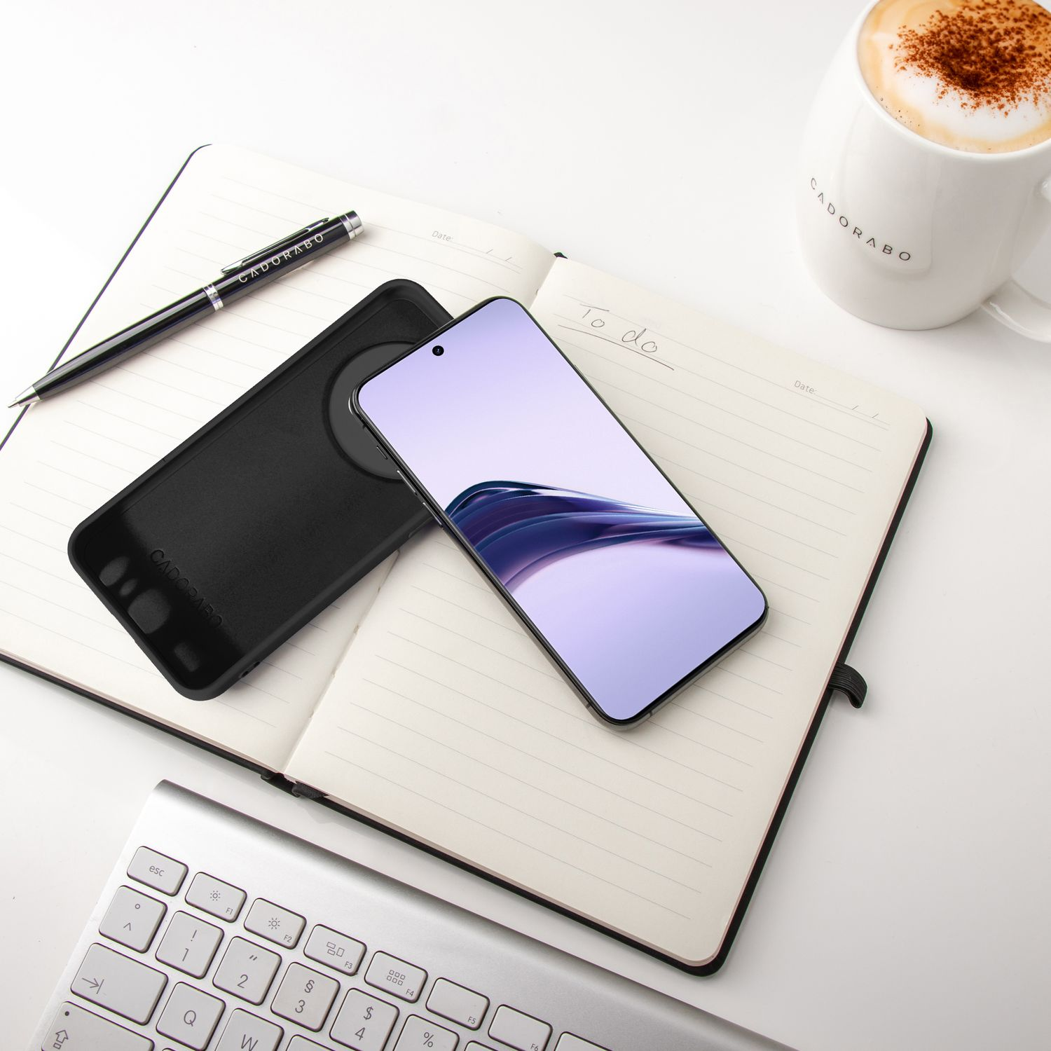 Telefon, Hülle, Stift und Kaffeetasse auf einem Notizbuch und einer Tastatur.