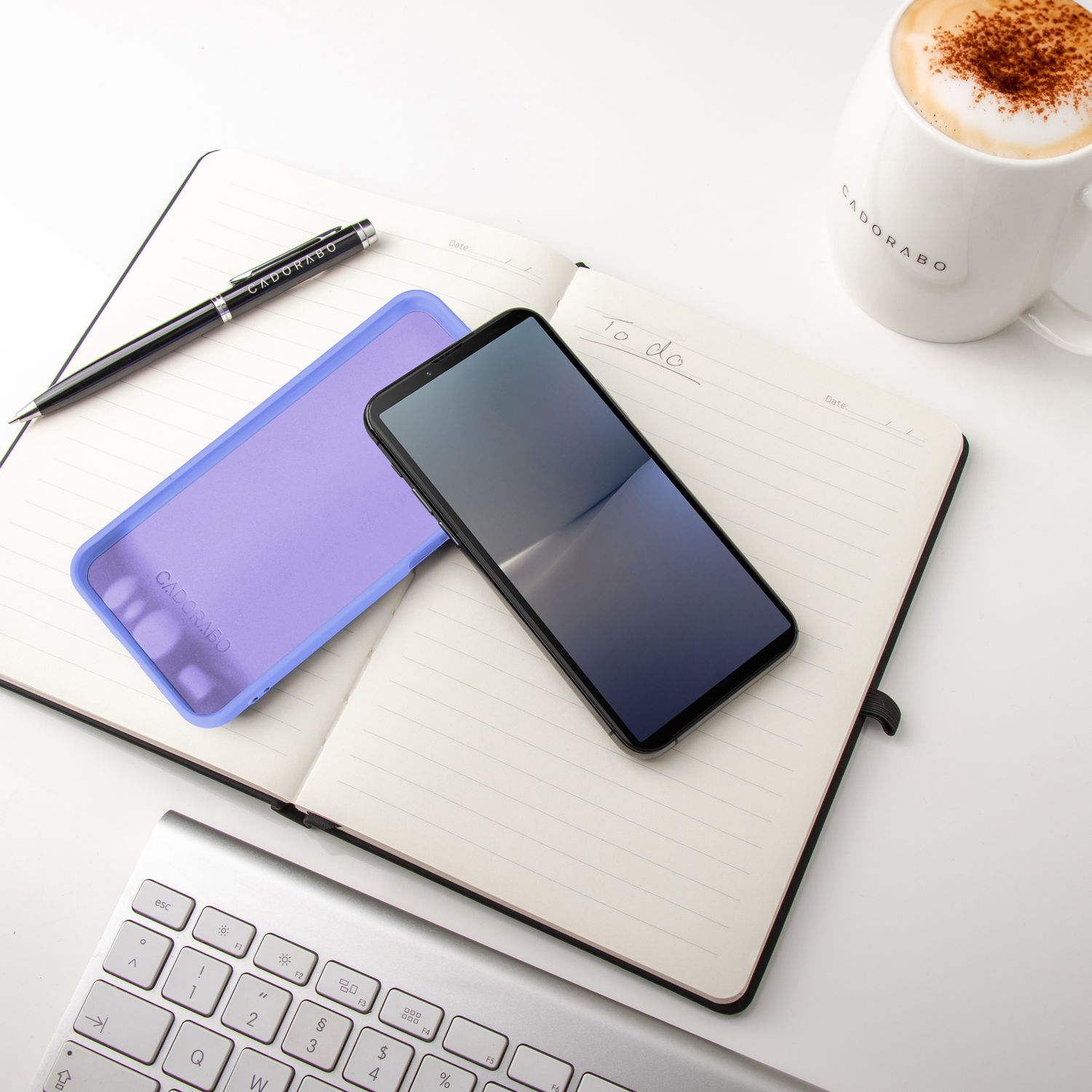 Notizbuch, Telefon, Stift und Becher auf weißer Oberfläche. Das Telefon hat einen leeren Bildschirm.
