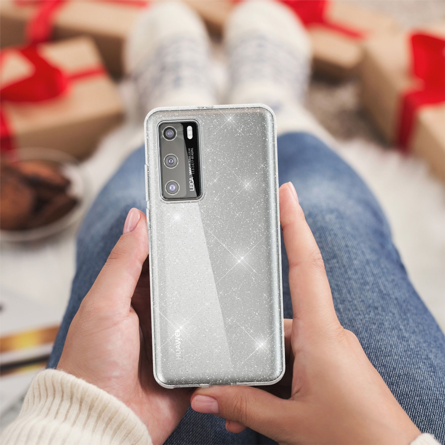 Hände halten eine glitzernde Handyhülle. Es hat drei Kameralinsen. Die Person sitzt mit Socken, mit Geschenken im Hintergrund.
