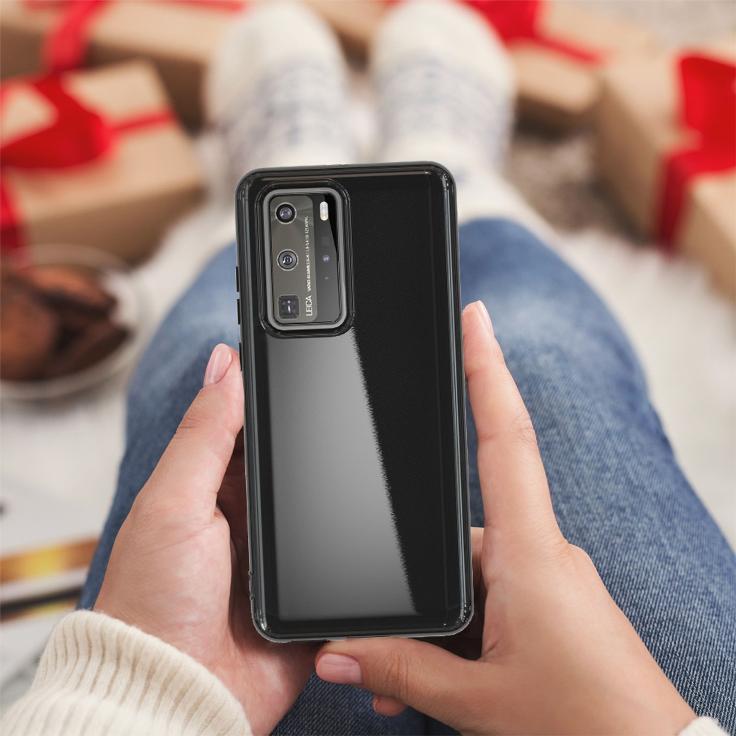 Hände halten ein schwarzes Telefon mit transparenter Hülle. Hintergrund: Beine mit Socken, Weihnachtsgeschenke.